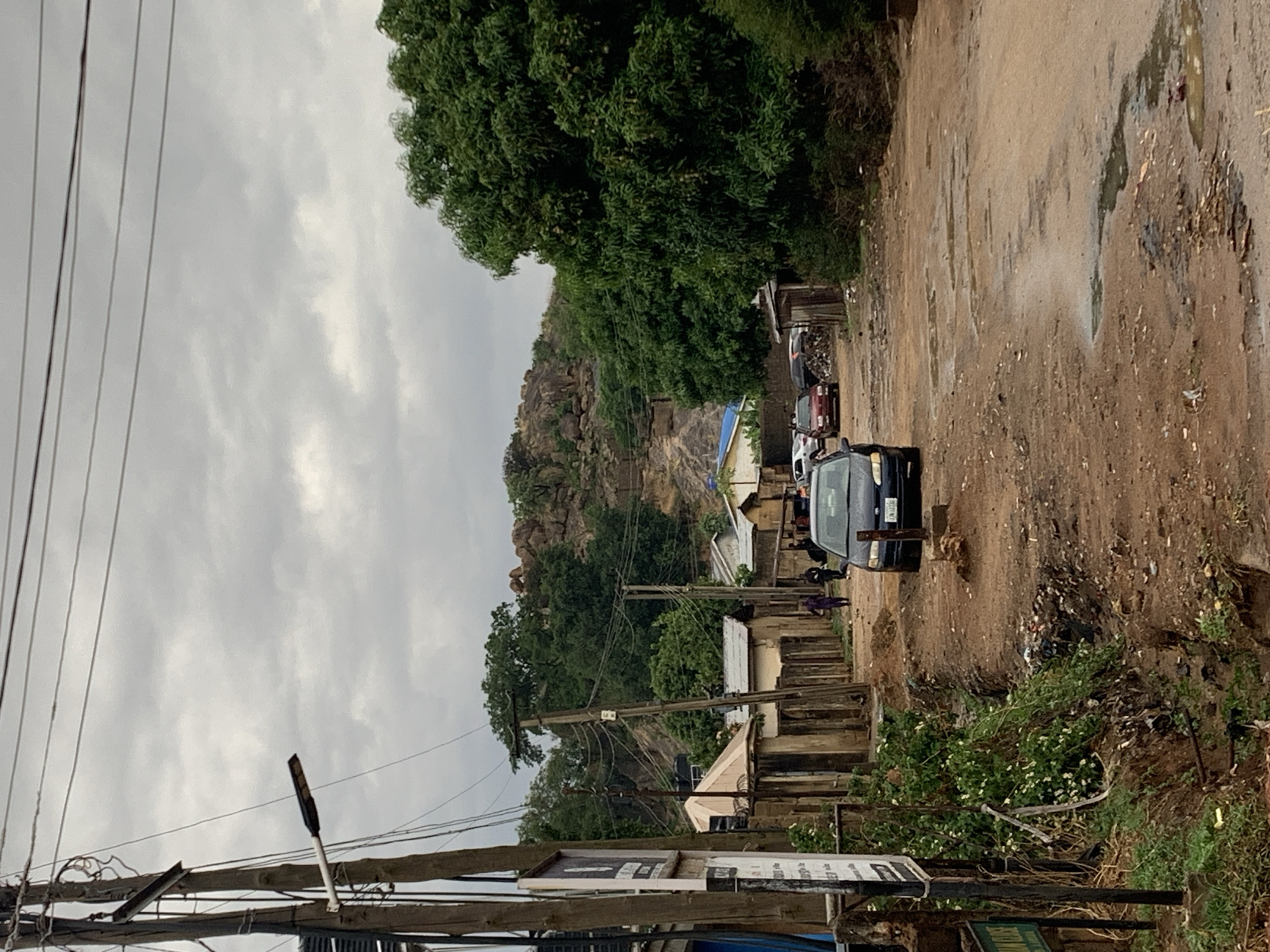 The image depicts a partially paved road in New Stadium, Nigeria, with a dark-colored car moving along it. The road surface is uneven, with patches of exposed dirt and standing water. To the right of the road, there is a buildup of trash and vegetation. Alongside the road are concrete structures, potentially walls or foundations of buildings. Power lines and a street lamp are visible overhead. In the background, a rocky hill can be seen, partially obscured by trees. Other vehicles are parked further up the road. Two people are walking along the street. The sky is overcast, creating a muted lighting in the scene.