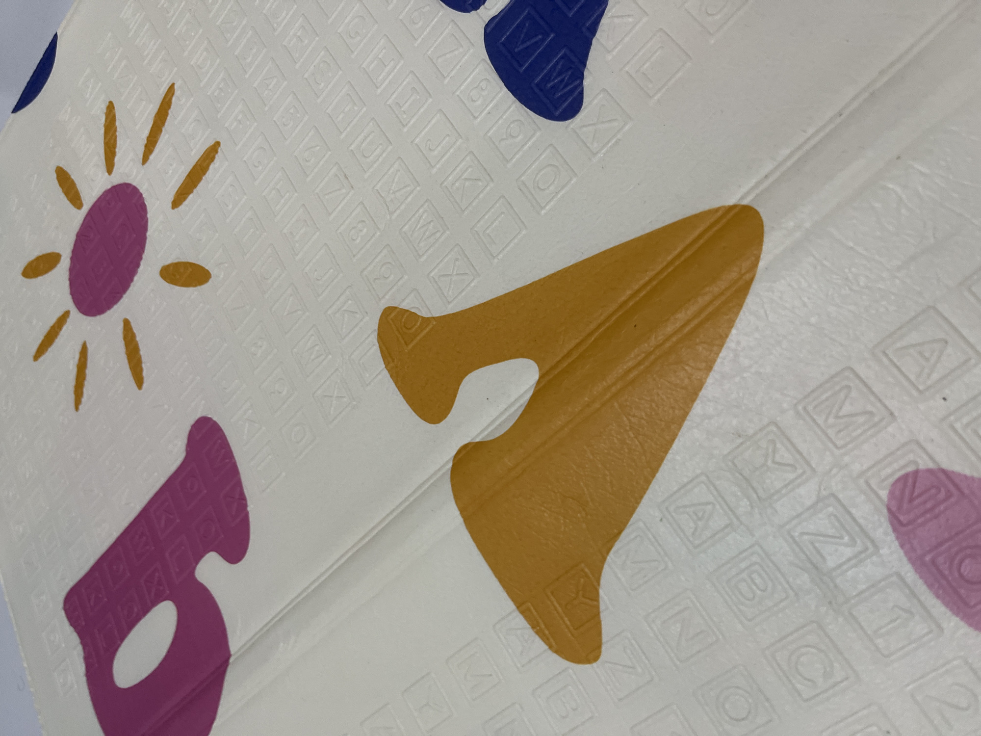 The image displays a close-up, downward-angled view of a white, textured surface, identified as a segment of a children's play mat, situated within the city of Lhokseumawe, Indonesia. The white substrate is patterned with a grid of debossed or embossed squares, each containing a single uppercase letter from the Latin alphabet or a numeral (0-9). Over this patterned base, larger, pigmented shapes are applied. In the upper-left quadrant, a magenta-colored letter 'A' is visible, partially cropped. In the upper-right, a design resembling a sun features a magenta central oval and radiating yellow, elongated oval rays. A large, marigold-yellow, curvilinear form occupies the lower-center and left portions of the frame, exhibiting a subtly textured surface. A small, dark blue, curvilinear element is present in the lower-right corner. A linear seam or fold is evident, traversing diagonally from the upper-center towards the lower-left of the image, segmenting the mat.