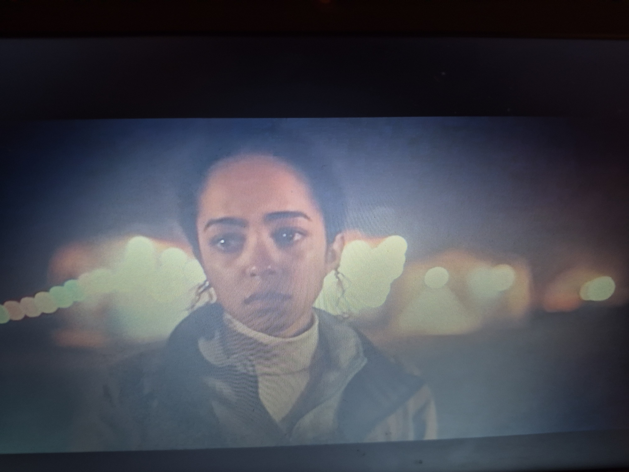 The image is a close-up shot of a young woman's face, appearing to be in distress. She has dark, curly hair and brown eyes. She is wearing a light-colored turtleneck sweater under a dark jacket. The background is blurred with out-of-focus lights, suggesting an outdoor setting at night, possibly in an urban environment. There are no discernible landmarks or features that would indicate a specific location within Sandwell, United Kingdom. The overall mood is somber and introspective.