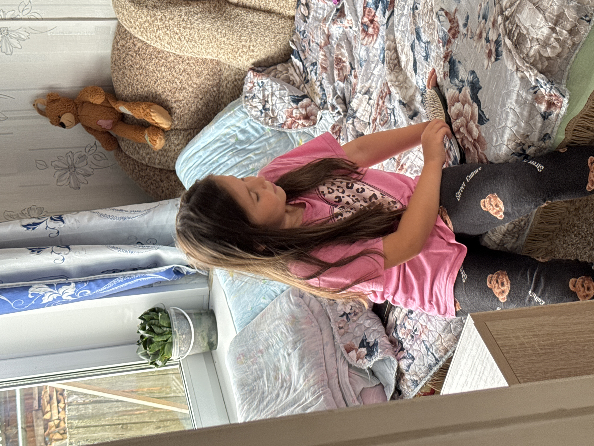 A young girl is lying on a bed, with a plush teddy bear beside her. She is wearing a pink t-shirt and dark leggings with teddy bear prints and the text "Street Culture" visible on them. The girl is holding a brush and appears to be brushing her long, brown hair. The bed is covered with a patterned comforter and a light blue blanket. In the background, a part of a couch or armchair is visible. The scene is indoors, near a window that looks out onto what appears to be a rustic outdoor area with wooden structures and stacked firewood. A small potted plant sits on the windowsill. The overall atmosphere suggests a casual, domestic setting, possibly during the daytime given the natural light coming from the window.