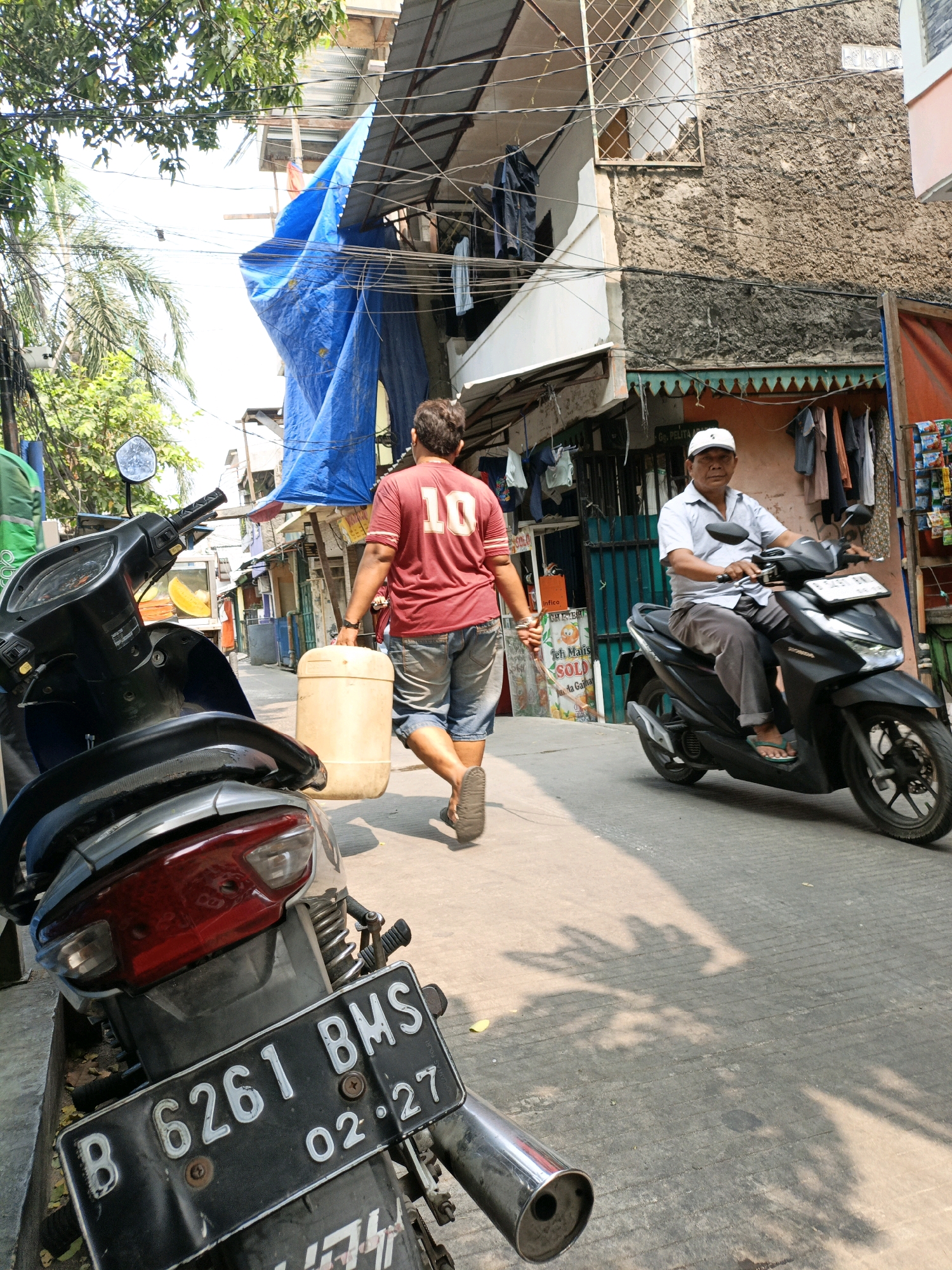 In West Jakarta, Indonesia, on a sunlit street, a person walks away from the camera, carrying a large, light-colored cylindrical container. The individual is wearing a red t-shirt with the number "10" on the back and denim shorts. To the right, a man wearing a white cap and a light blue collared shirt rides a black motorcycle. The street is lined with buildings, some of which appear to be shops with goods displayed outside. A parked motorcycle is visible in the foreground on the left, its license plate reading "B 6261 BMS 02-27". Overhanging wires and a blue tarp are present above the street, suggesting a densely populated urban environment. The ground is paved with concrete and shows shadows cast by overhead objects.