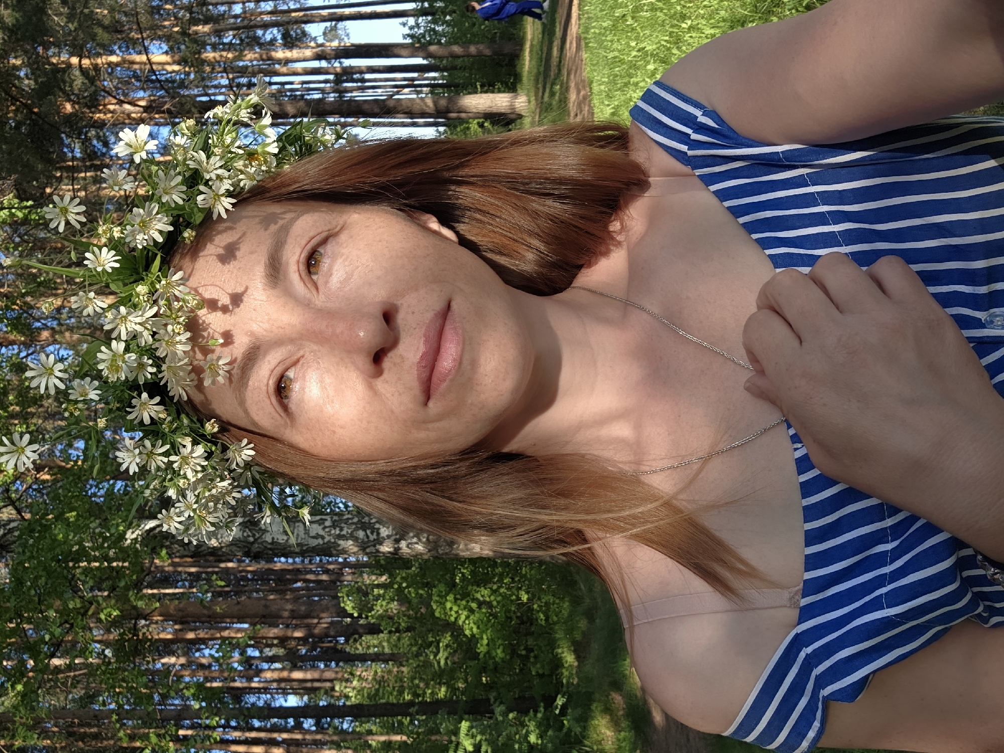 A woman with light brown hair and a flower crown is centered in the image. She is wearing a blue and white striped top, with the forest visible in the background. The location is Chaikovsky, Russia.