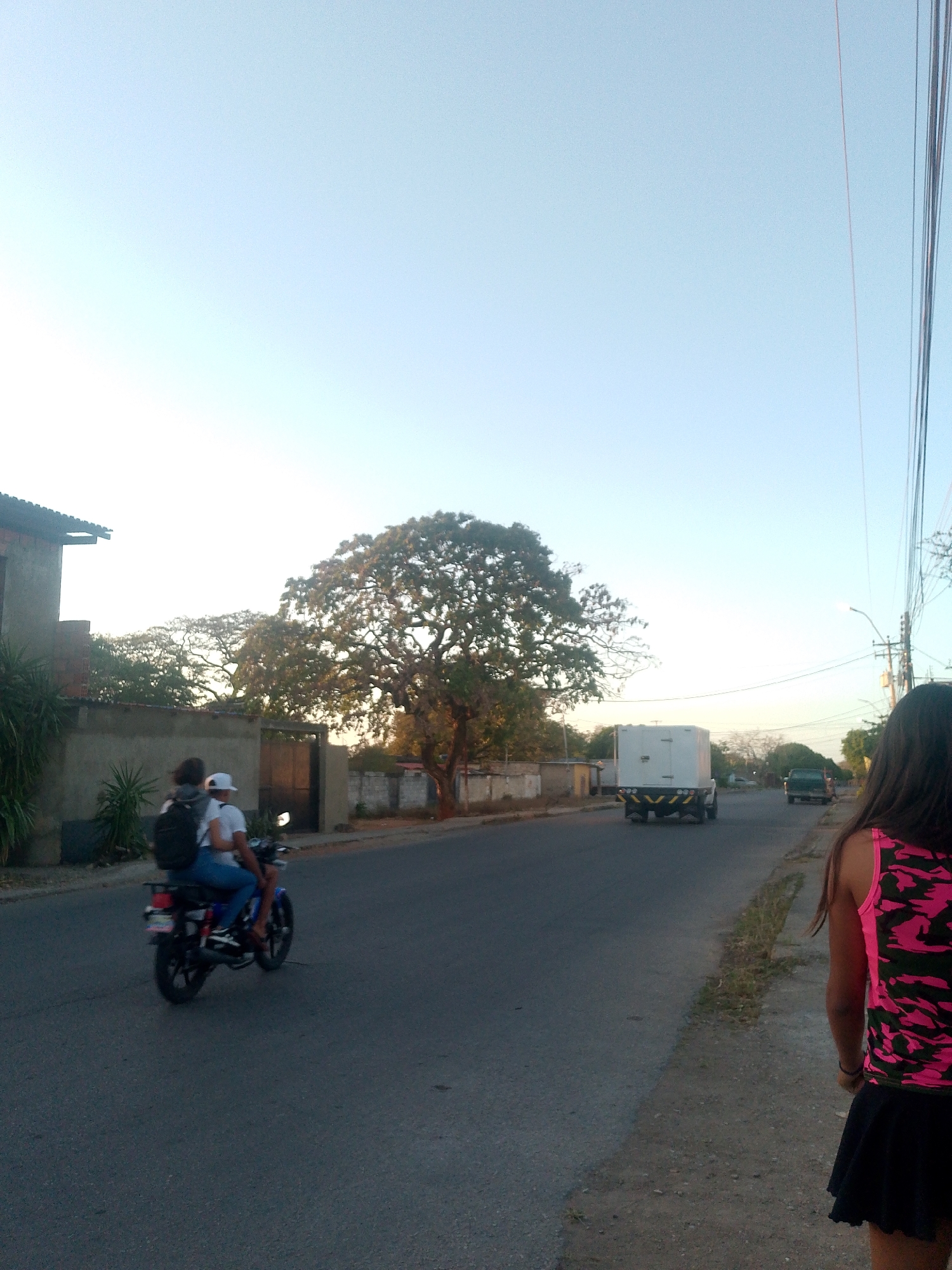 The image depicts a street scene in what appears to be Juan Griego, Venezuela. The sky is light blue, suggesting either early morning or late afternoon. A two-lane asphalt road runs through the scene.
On the left side of the road, a motorcycle with two people is traveling away from the camera. The driver wears a white shirt and a white cap. The passenger, seated behind, is wearing a dark backpack.
A beige concrete wall borders the left side of the road, with a building partially visible behind it. In the distance, a large tree dominates the view.
On the right side of the road, a white box truck is also driving away from the camera. Further down the road, a dark-colored SUV is visible.
A girl wearing a black skirt and a pink and black camouflage tank top stands on the right side of the road, facing away from the camera. The ground to her right appears to be a grassy verge. Power lines are visible above on the right side of the image.