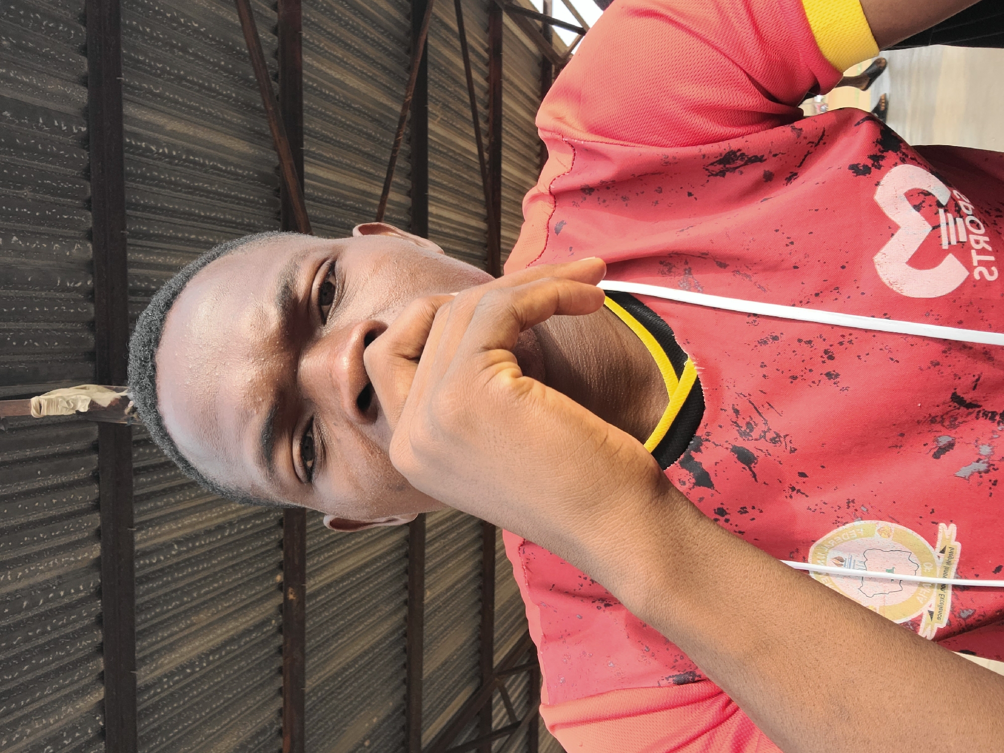 A young, dark-skinned man wearing a red shirt is holding his hand near his face and looking at the camera. The shirt has yellow and black accents on the neckline and sleeves, and a logo on the chest. The background shows corrugated metal siding and structural supports, suggesting an outdoor shelter. The man is likely located in Jos, Nigeria.