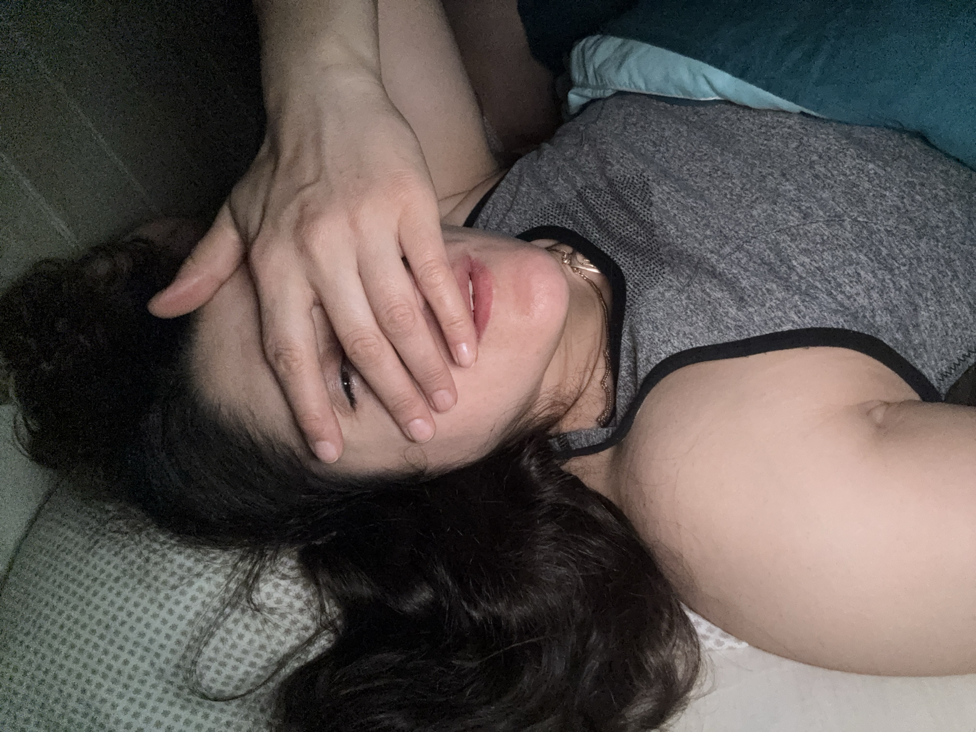 A woman lies on a patterned pillow in a dimly lit room, partially covered by a blue blanket. She wears a gray tank top with black trim. Her dark hair is spread across the pillow. Her hand covers a portion of her face, but one eye is visible. The woman's location is not identified in the image. The location is not Tobolsk, Russia.