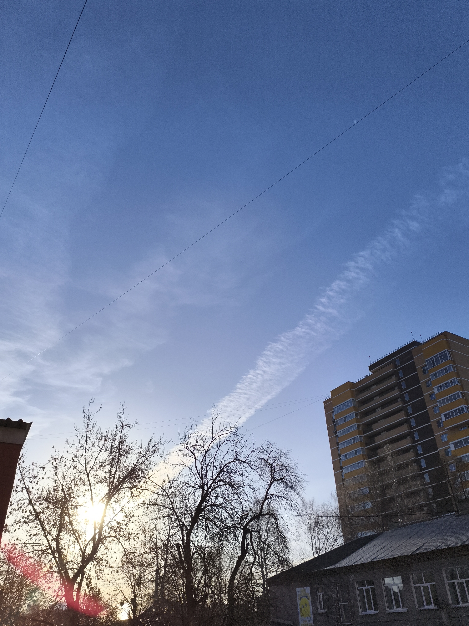 The image depicts a view upwards toward a mostly clear blue sky with some wispy clouds. A contrail stretches diagonally across the sky from the lower left towards the upper right, terminating near a tall yellow and brown building. Two power lines cross the upper portion of the frame.
In the lower portion of the image, there are bare trees and buildings. The sun is shining through the branches of one tree, creating a bright spot and lens flare. A portion of a brick building is visible on the left, and a grey building with white-framed windows is below the apartment building.