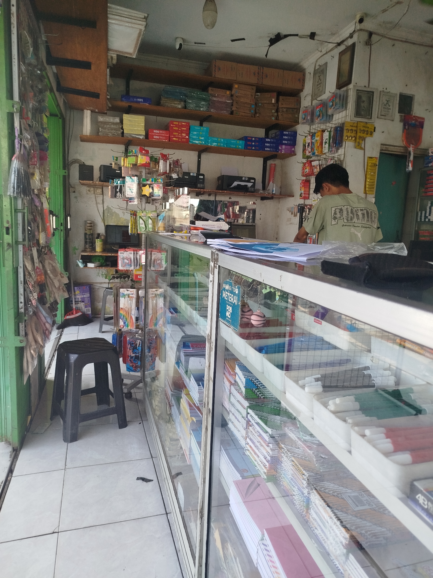 The image shows the interior of a small convenience store, likely a "warung" or small shop, located in Mranggen, Indonesia. The shop sells a variety of goods including stationery, office supplies, and possibly some food items.
In the foreground, a glass display counter is filled with items such as notebooks, pens, and other stationery supplies. Some of these items are organized in white plastic trays, with pens of different colors visible. The counter is lit from above, illuminating the products.
Behind the counter, a person, presumably the shopkeeper, is partially visible. They are wearing a light-colored t-shirt with a graphic print and a dark baseball cap, and they are facing away from the camera, attending to something behind the counter.
The shop is packed with merchandise, with shelves lining the walls above and behind the counter. These shelves are stocked with boxes of paper, stationery packs, and various other retail items. To the left of the frame, an entryway is visible with green metal folding doors, through which more items are displayed hanging on racks.
The environment appears to be a typical street-level shop in Indonesia. The walls are painted a light color, and the floor is tiled. A black plastic stool is placed in front of the counter. Various framed pictures and advertisements are mounted on the walls. The overall impression is of a busy, well-stocked small business.