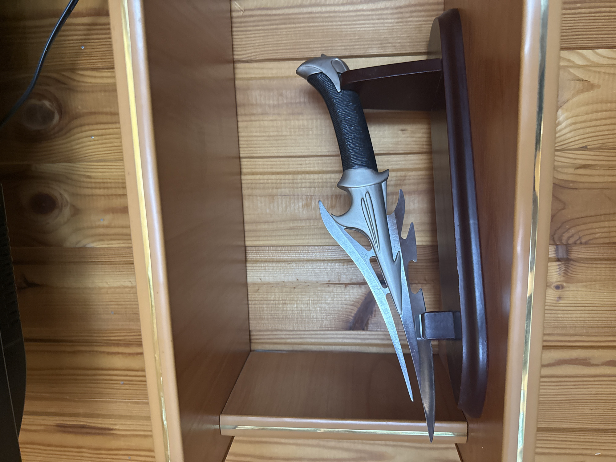 A decorative dagger with a black, textured handle and a silver, pointed blade is displayed upright in a wooden shelf. The dagger is attached to a dark brown wooden mount, which is secured to the back wall of the shelf. The shelf itself is made of light-colored wood with horizontal grain. The back wall of the shelf is also made of wood with horizontal planks. The shelf is open at the front, and the bottom of the shelf is visible. A dark electrical cord is partially visible on the left side of the image, extending from the top left corner. The lighting suggests an indoor setting, likely during the daytime, with natural light illuminating the scene. There is no discernible text in the image. The scene is static, with the dagger presented as an exhibit.