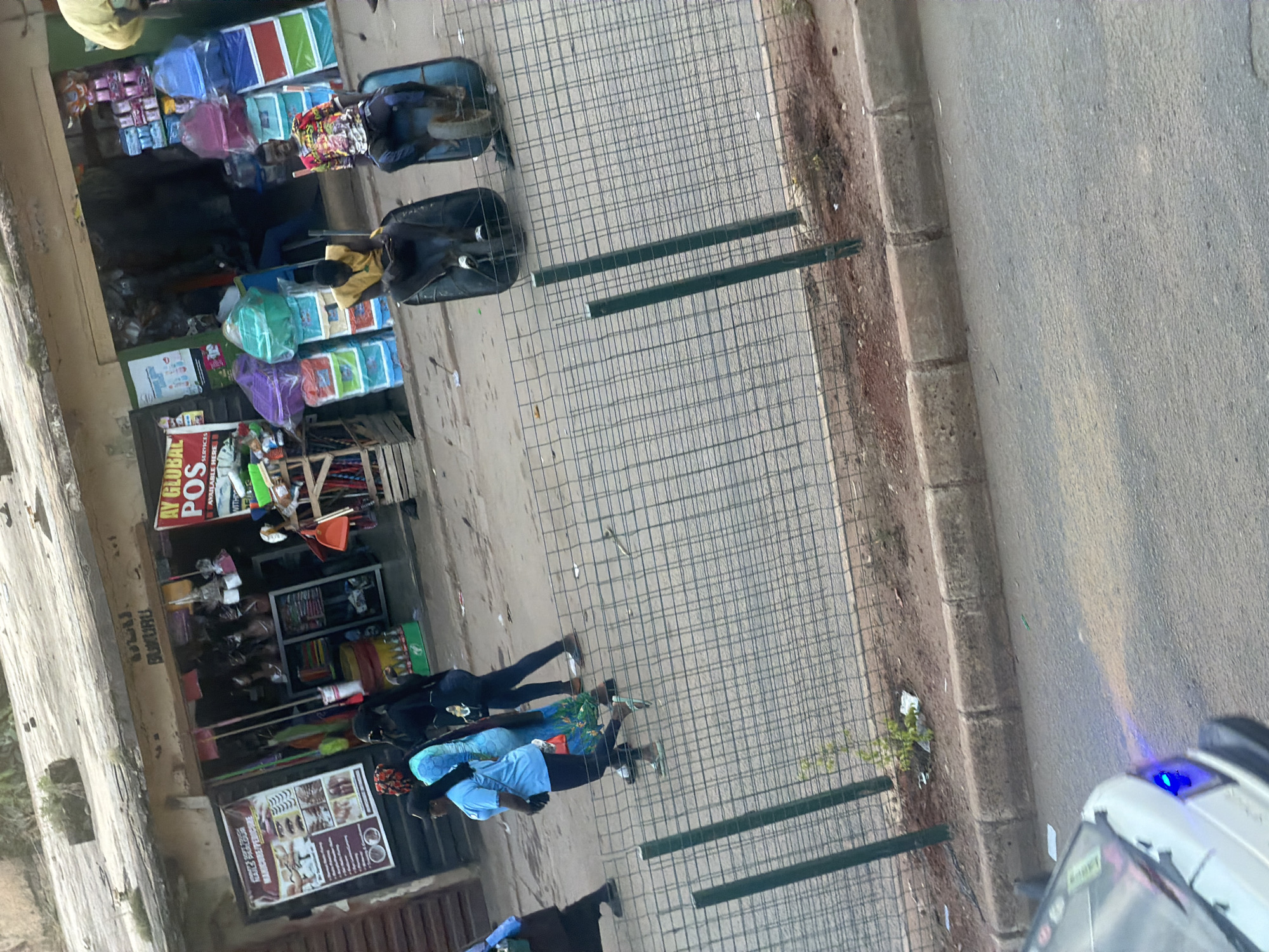 The image depicts a street scene in Bukuru, Nigeria, in front of a shop. The shop has a weathered awning and displays various goods, including wigs, clothing, and what appear to be storage containers. Two men are seated in wheelbarrows, one in a yellow shirt and the other in a colorful patterned shirt. Several women are walking on the sidewalk, one in a blue shirt and another in a blue and green patterned dress. A white vehicle with blue headlights is visible in the lower left corner, partially obscuring the foreground. A wire fence runs along the sidewalk, separating it from the street. The ground is paved with asphalt and cobblestone. The overall scene suggests a busy commercial area.