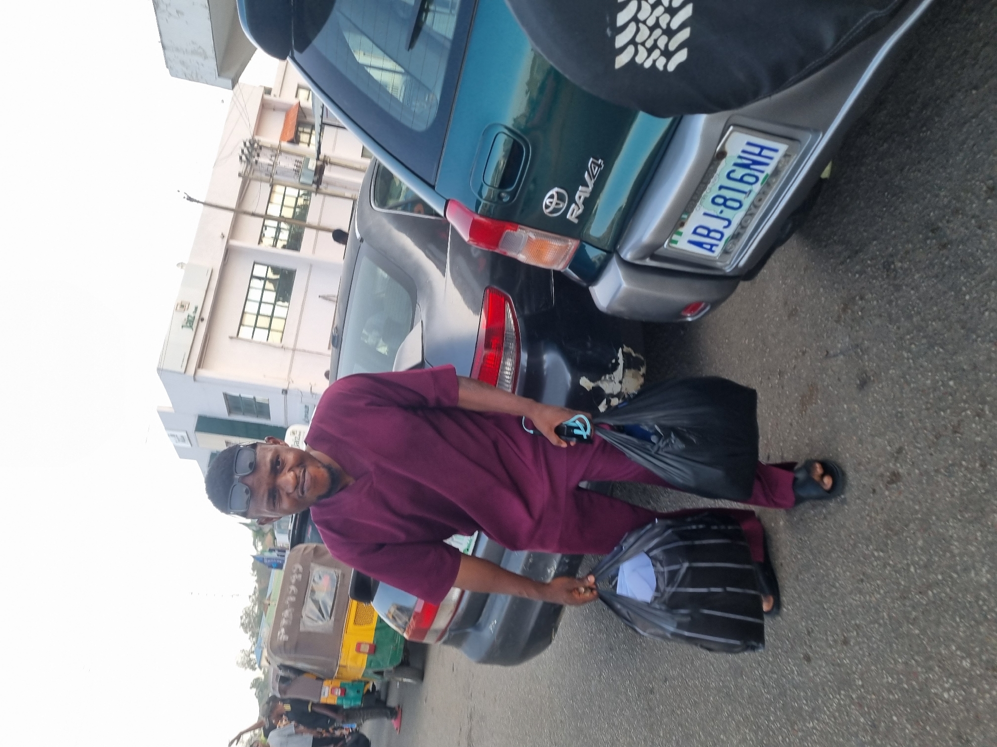 A man wearing a maroon outfit, glasses, and sandals stands on a street in Jos, Nigeria. He holds two black plastic bags. Behind him are several vehicles including a green Toyota RAV4 with license plate ABJ-816NH, a black car, and a yellow and green auto rickshaw. Other people are visible in the background. A building with windows and an electrical pole can be seen across the street. The image is taken during daylight.