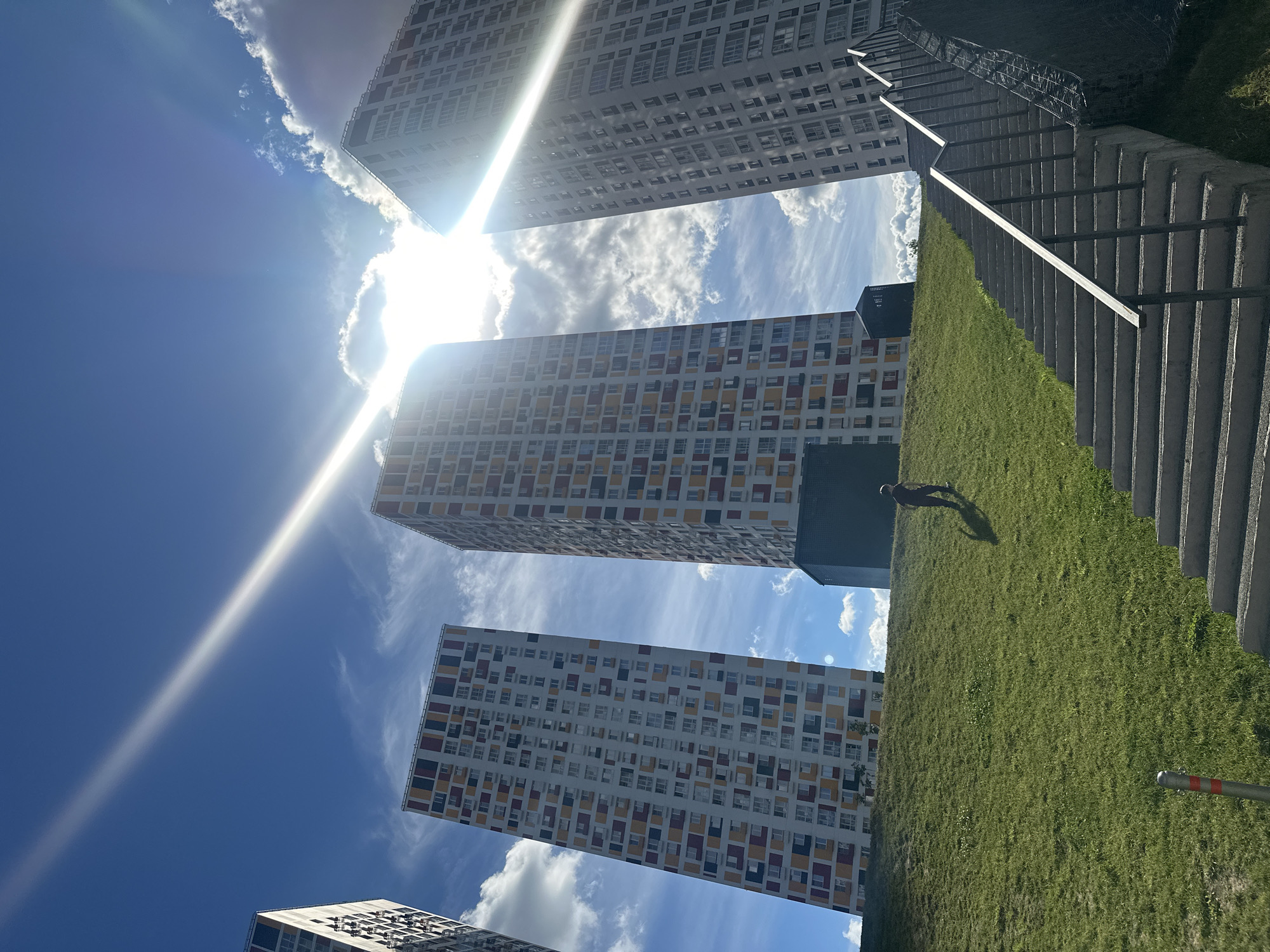 The image shows a low-angle view of several tall, modern apartment buildings against a bright blue sky with scattered clouds. The sun is intensely bright, causing lens flare and washing out parts of the sky. The buildings are multi-story structures with repetitive window patterns. One building in the foreground has a facade with colored panels in yellow, red, and dark blue interspersed with white.
To the right of the buildings, there is a grassy slope leading up to a concrete staircase with a metal railing. A lone figure is visible on the grassy slope, casting a shadow, appearing to be walking. The scene suggests daytime, likely a sunny afternoon, given the strong sunlight and clear sky. The setting appears to be an urban residential area, possibly a park or courtyard between buildings, in Khimki, Russia, as indicated by the location context. The overall impression is one of modern urban architecture and a bright, sunny day.