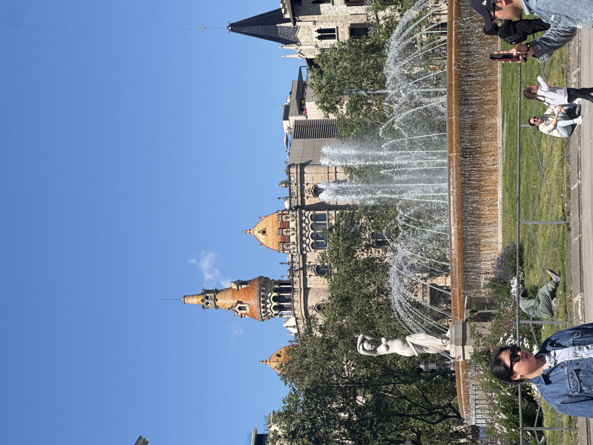 The image captures a scene in Barcelona, Spain, featuring a fountain and the architecture of Castell dels Tres Dragons (Castle of the Three Dragons), a building in the Parc de la Ciutadella. The building's spires, towers, and facade are visible behind the fountain. The fountain features water jets and sculptures. In the foreground, people are interacting with the scene: some are taking pictures, while others are sitting nearby. The weather appears sunny, with a clear blue sky visible above. The overall impression is a vibrant urban scene, combining historical architecture with public space and human activity.