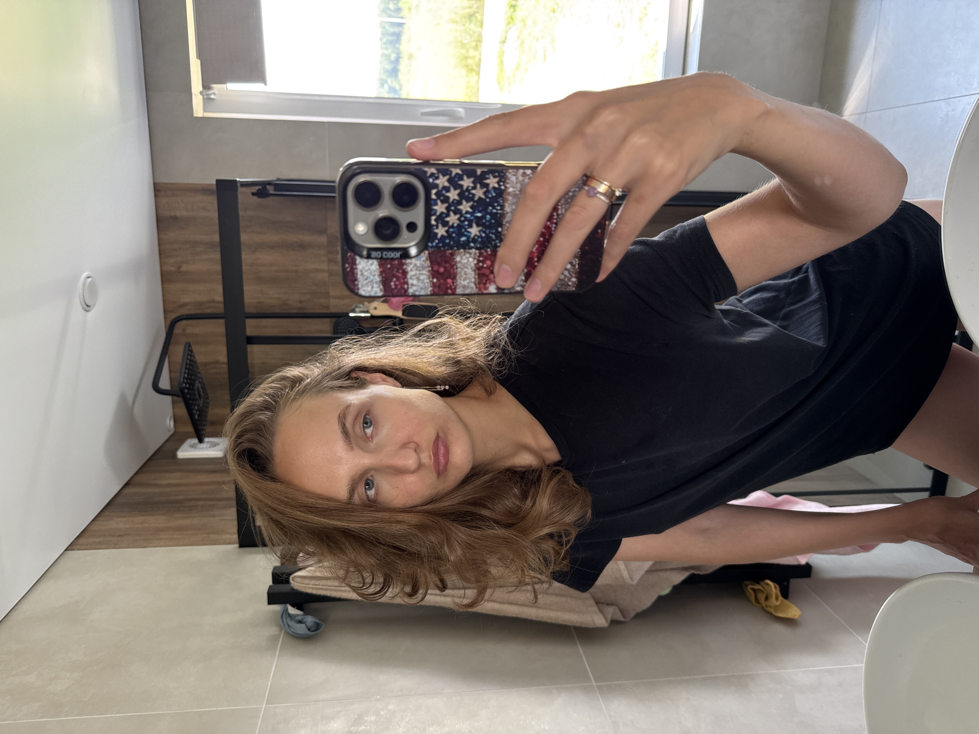 A woman with brown hair is taking a mirror selfie in a modern bathroom. She is wearing a black t-shirt and dark shorts. Her hair is slightly messy and falls around her shoulders. She is holding up a phone with a glittery American flag case. In the background, there is a shower, a window with natural light, and tiled walls. There are also towels hanging on a rack. The location is specified as Podshivalovo, Russia.