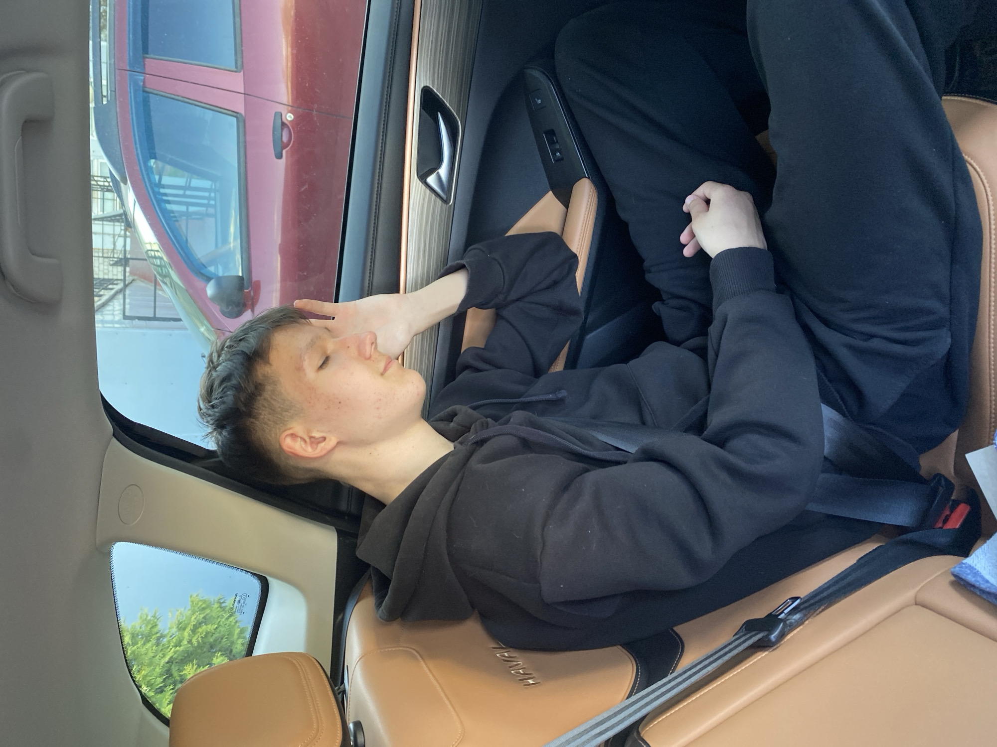 A young male is reclined in a tan leather car seat with black accents, wearing black sweatpants and a black hooded sweatshirt. He has a seatbelt fastened across him. His right hand is near his face. The interior of the car includes a wood-grain-like panel on the door. The exterior visible through the car windows includes a red vehicle and foliage. There's no information to suggest the location is specifically Chaikovsky, Russia. The text "HAVAL" is visible on the seat.