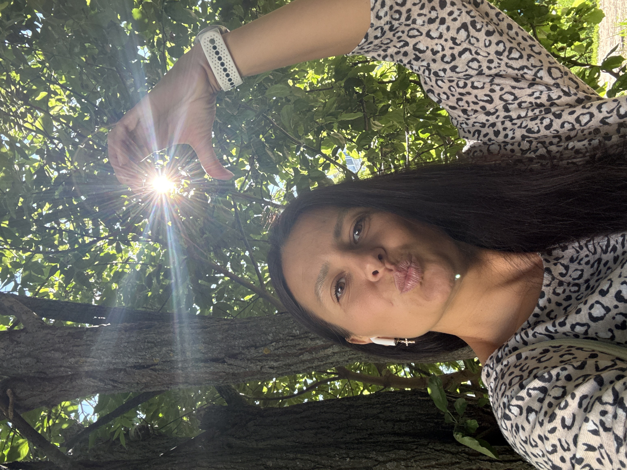 A woman with dark hair, wearing a leopard-print shirt and a white fitness tracker on her left wrist, is holding her hand up to form a heart shape around the sun. She is looking at the camera with her lips pursed. She is wearing white earbuds, one with a cross charm. The background is out of focus and shows trees and foliage, with sunlight streaming through the leaves. The image appears to have been taken outdoors. The location is identified as Chaikovsky, Russia.