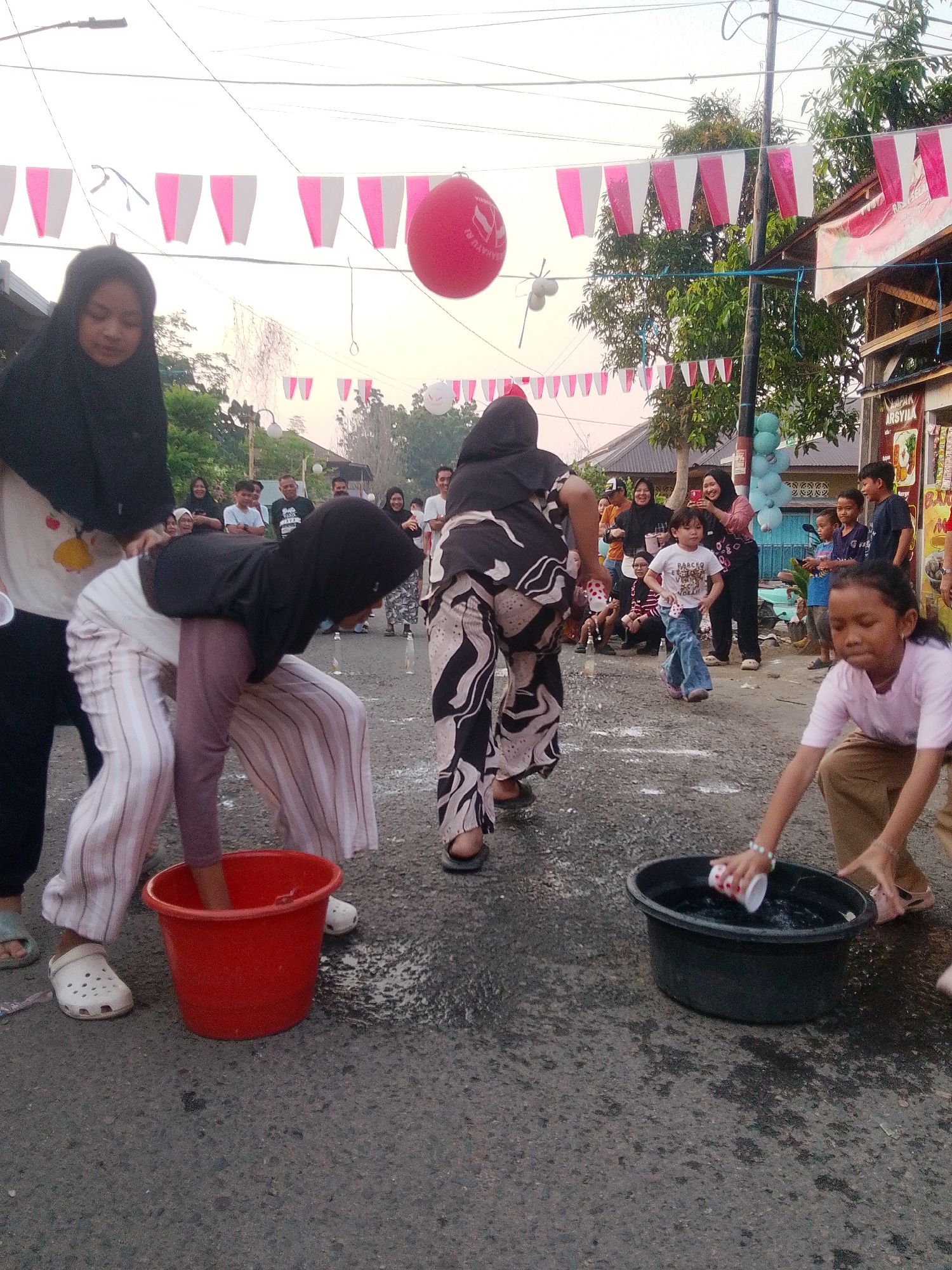In Barabai, Indonesia, a street event is underway with children and adults participating in a game. A young girl, wearing a light pink t-shirt and brown pants, is shown squatting down and attempting to fill a white and red cup from a black basin filled with water. To her right, another child, a boy wearing a white t-shirt and blue jeans, is running. In the background, a group of people are watching the game, some of whom are wearing traditional Islamic head coverings. Decorations of pink and white flags are strung across the street, and a large red balloon floats in the sky, indicating a festive atmosphere. The ground is wet, suggesting water has been spilled during the game. The scene appears to be taking place during daylight hours, with soft lighting suggesting it may be late afternoon or early evening.