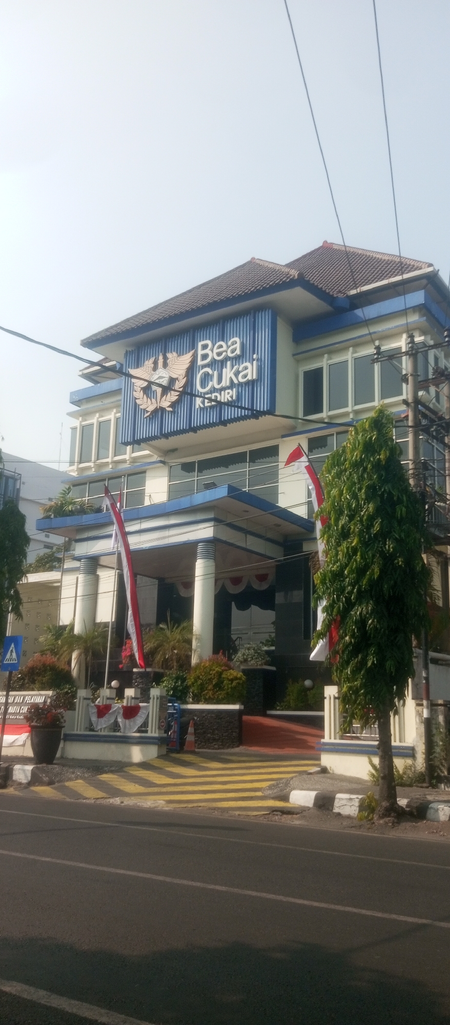 The image shows the exterior of the "Bea Cukai" building in Kediri City, Indonesia. The building is a multi-story structure with a blue and white facade. The words "Bea Cukai" and "KEDIRI" are displayed prominently on a sign above the entrance. Indonesian flags are visible, as well as some greenery and a tree in front of the building. The foreground shows a street with yellow and white pedestrian crossing markings. The overall scene depicts a government office building in an urban setting on a clear day.