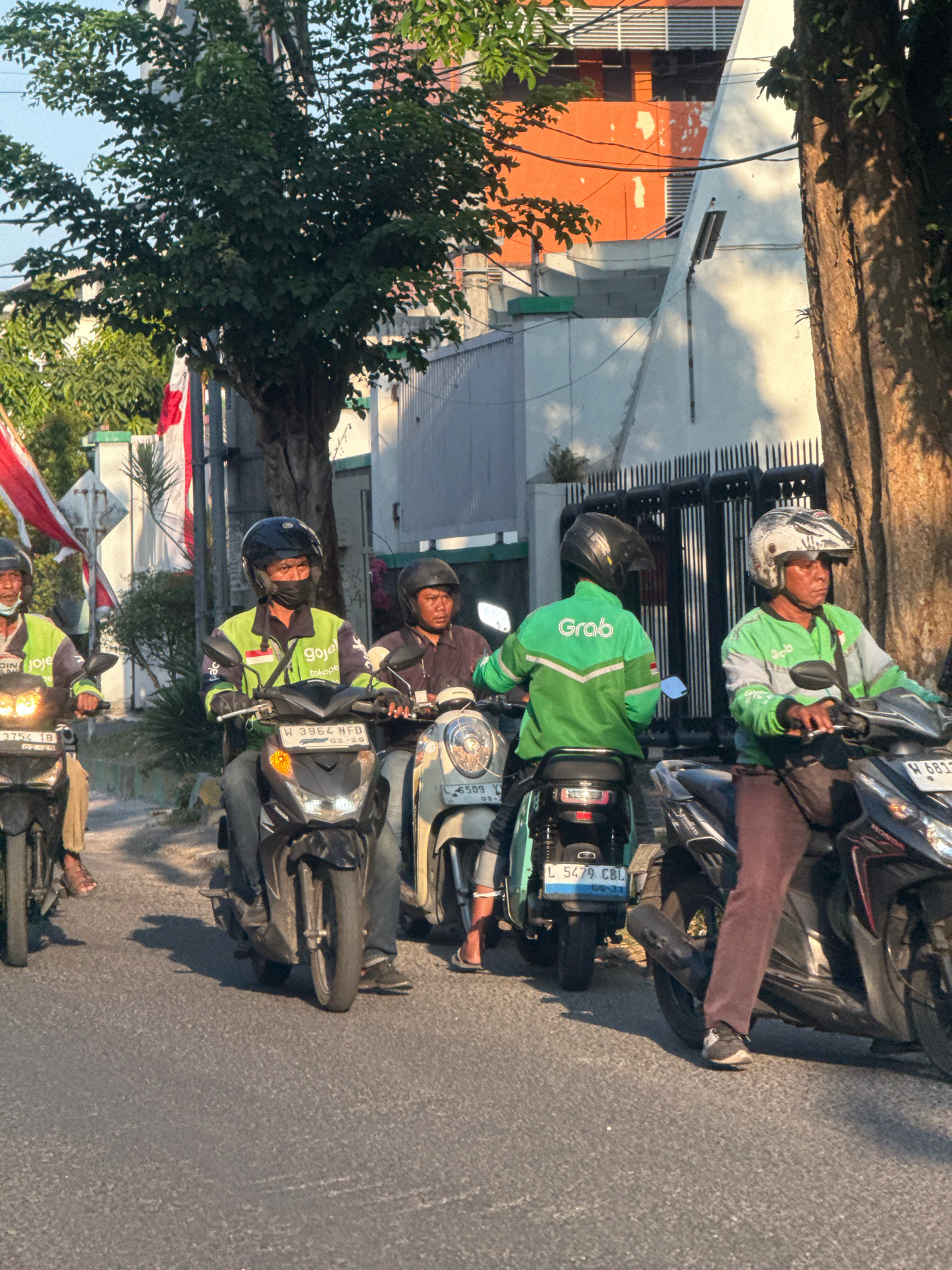 The image shows four motorcycle riders on a street in Surabaya, Indonesia. Three of the riders are wearing green jackets, indicating they are likely delivery or ride-sharing drivers for services like Gojek or Grab. The rider in the center is wearing a black face mask and a green and yellow Gojek jacket. The rider to his right is wearing a white and green jacket with a Gojek logo. The rider to the far left is wearing a green jacket with a Gojek logo, and his motorcycle has "Gojek" written on it. The rider on the far right is wearing a green jacket with "Grab" written on it. The motorcycles are parked or moving slowly in traffic, and the riders are looking forward. In the background, there are buildings, trees, and Indonesian flags. The sun is shining, casting shadows on the road. The scene depicts a typical urban street environment in Indonesia.