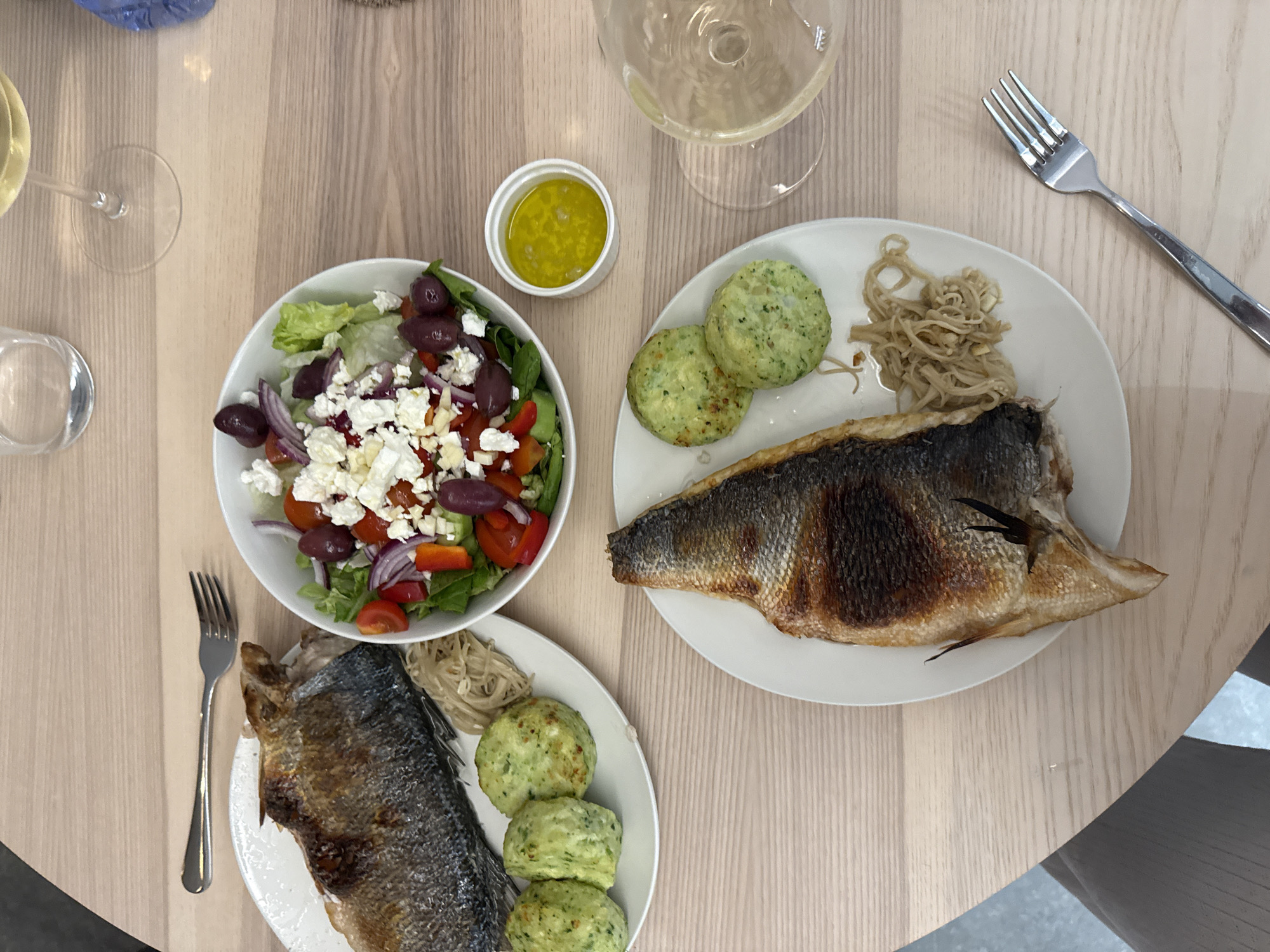 The image depicts a dining table with two plates of food, a salad, a small dish of dressing, two wine glasses, and two forks. The food on each plate consists of a whole grilled fish, a small pile of noodles, and two or three green savory cakes. The salad is a Greek salad with lettuce, tomatoes, red onion, olives, and feta cheese. The dressing is a yellow-green liquid. The location may be a restaurant in Livadia Larnakas, Cyprus.