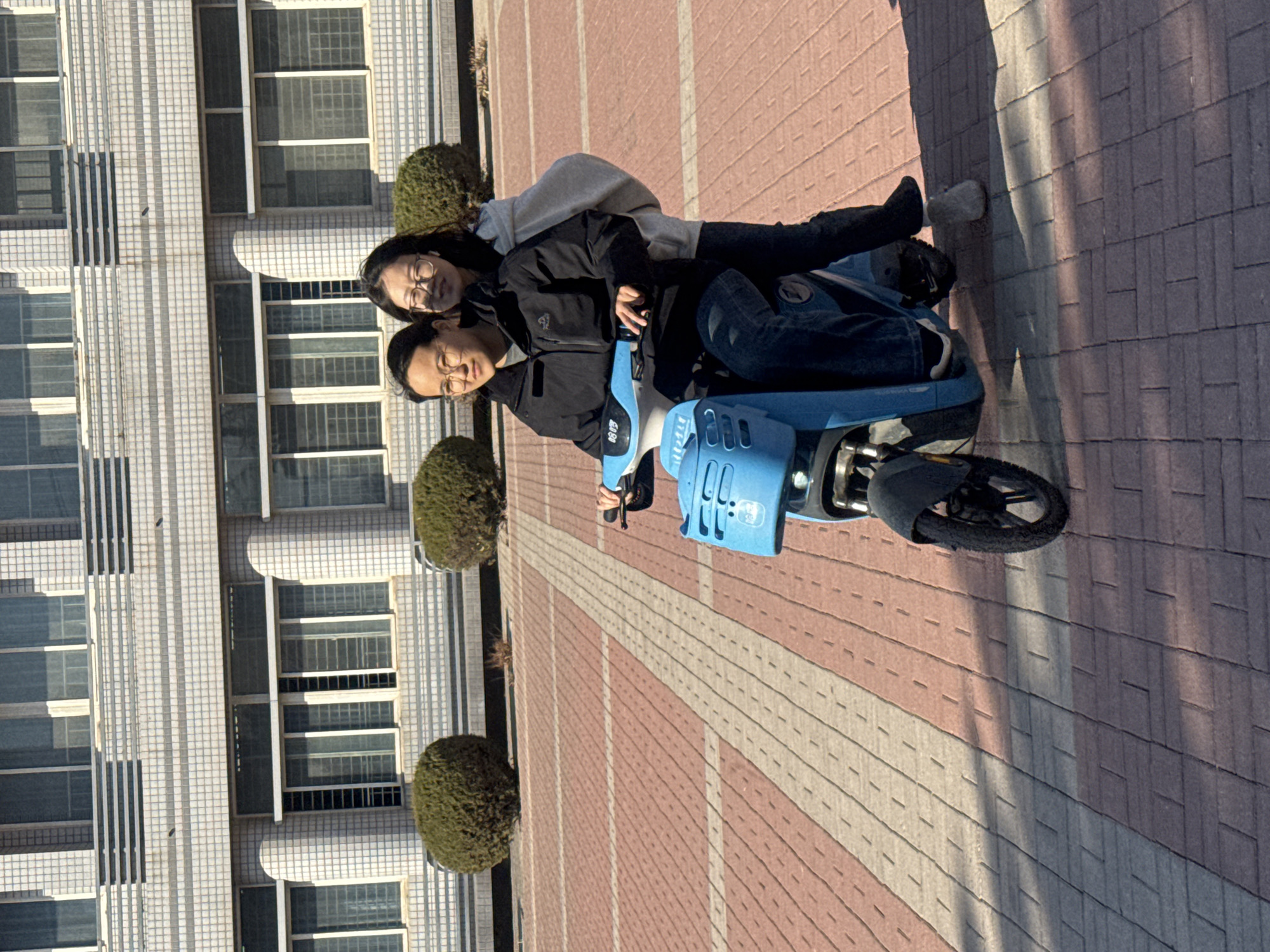 Two East Asian females are positioned on a bright blue electric scooter on a paved outdoor surface in Zhongtun, China. The scooter faces slightly right of the camera. The woman in the driver's seat, wearing glasses, a black padded jacket, dark jeans, and white-soled shoes, holds the handlebars. The woman in the passenger seat, also wearing glasses, a light grey hooded sweatshirt, dark pants, and light grey slippers, sits behind the driver. Both women are looking towards the camera, with the passenger exhibiting a slight smile. The scooter has a front basket and a digital display showing "0805." Behind them, a large, multi-story building with a light-colored tiled facade and numerous rectangular windows extends across the background. Several spherical, dark green shrubs are visible between the subjects and the building. The ground is paved with reddish-brown interlocking pavers, interspersed with lighter beige strips. Strong sunlight illuminates the scene, casting distinct shadows, including a large shadow from an unseen object in the lower left foreground.