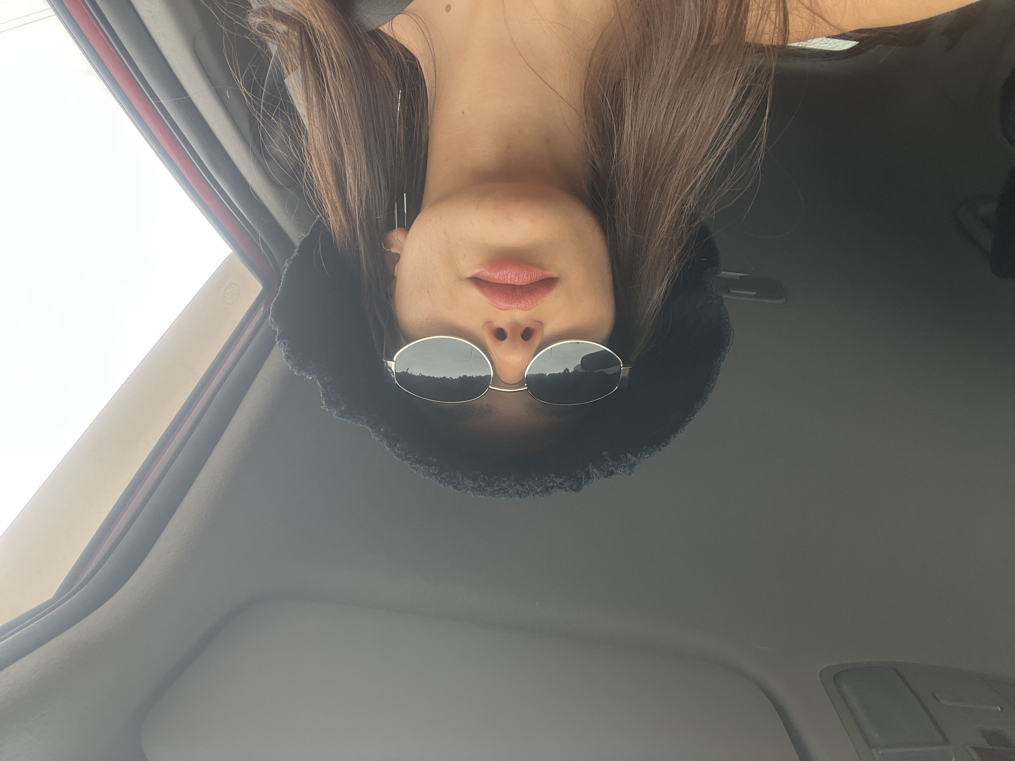 The media file presents a low-angle, close-up photograph of a person, likely female, inside a car. The person is positioned upside down in the original image. She has long, dark brown hair and is wearing a dark, possibly fuzzy, bucket hat along with round, reflective sunglasses that cover her eyes. Her lips are visible, appearing to have a pink or nude lip color. A small portion of her neck and shoulder is also visible.
The setting is the interior of a car, with the beige/grey headliner and an overhead console visible in the background. A bright, overcast sky can be seen through the car window on the left, which also shows a red trim on the window frame. The reflective sunglasses mirror an outdoor landscape, possibly trees or hills, under the bright sky. A faint circular emblem containing the number "62" is visible on the car window. The ample natural light indicates the photo was taken during the daytime. The image appears to be a selfie or a portrait taken from a unique angle within the vehicle, located in Shmidtovka, Russia.