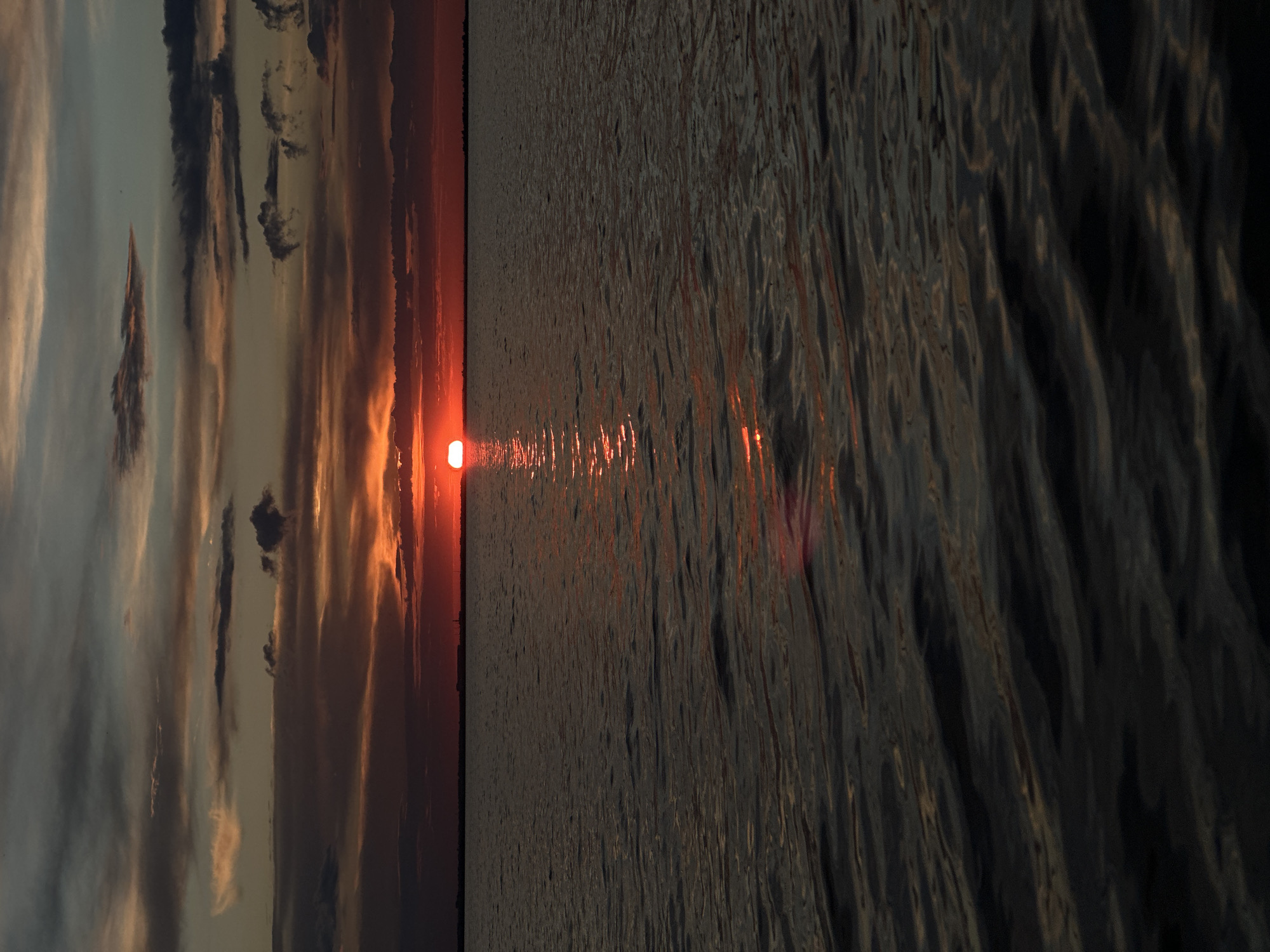 The image shows a sunset over a body of water. The sun is a bright orange disc just above the horizon, casting a shimmering reflection on the dark water. The sky is filled with dramatic clouds in shades of orange, red, and gray. The horizon line is straight and appears to be the edge of land, with faint silhouettes of structures visible on the far left. The foreground consists of rippling water, dark and almost black, with subtle hints of the sunset's colors reflected on its surface. There are no people or animals visible in the image. The scene conveys a sense of calm and natural beauty.