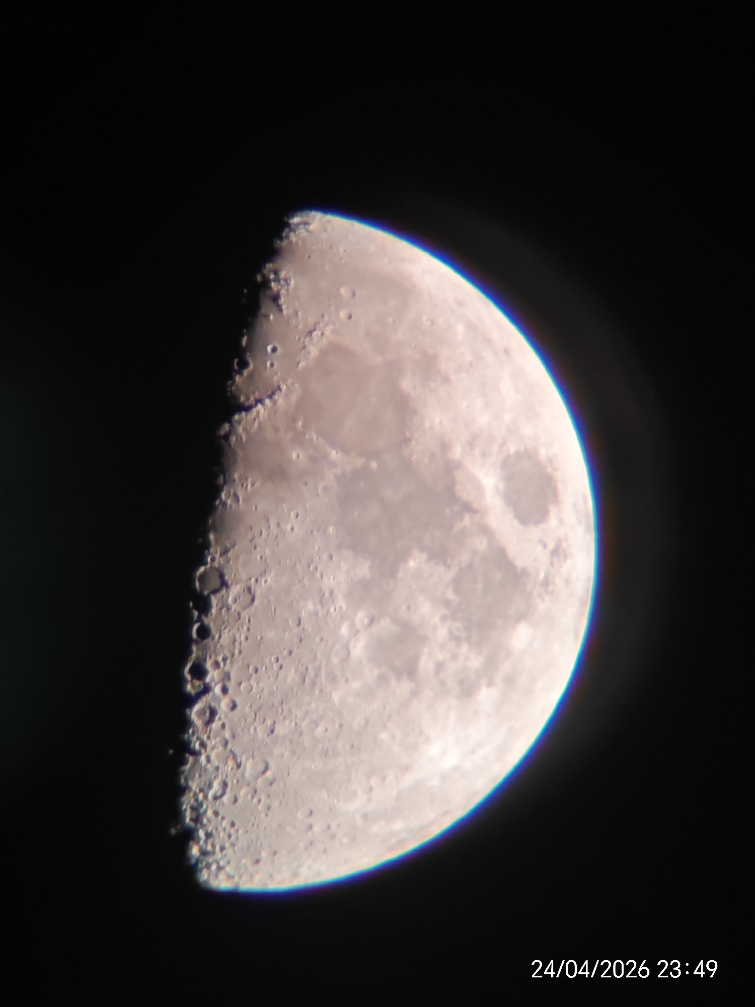 A clear photograph showcases the Moon in a half-illuminated phase, likely a first or last quarter, against a dark, featureless background of space. The illuminated portion of the lunar surface reveals intricate details, including numerous craters of varying sizes, particularly prominent along the rugged terminator line where shadows cast by crater rims enhance their depth and definition. Large, relatively smoother, and darker areas, indicative of lunar maria, are also visible.
A notable optical artifact, chromatic aberration, is evident as a blue and purple fringe along the brightly lit outer edge of the Moon, particularly noticeable on the right side. The overall clarity of the image suggests it was captured with a telescope or high-magnification lens.
In the bottom right corner of the image, a timestamp is present, reading "24/04/2026 23:49," indicating the date and time the photograph was taken. No other objects, people, or discernible landscape features are visible, emphasizing the Moon as the sole subject.