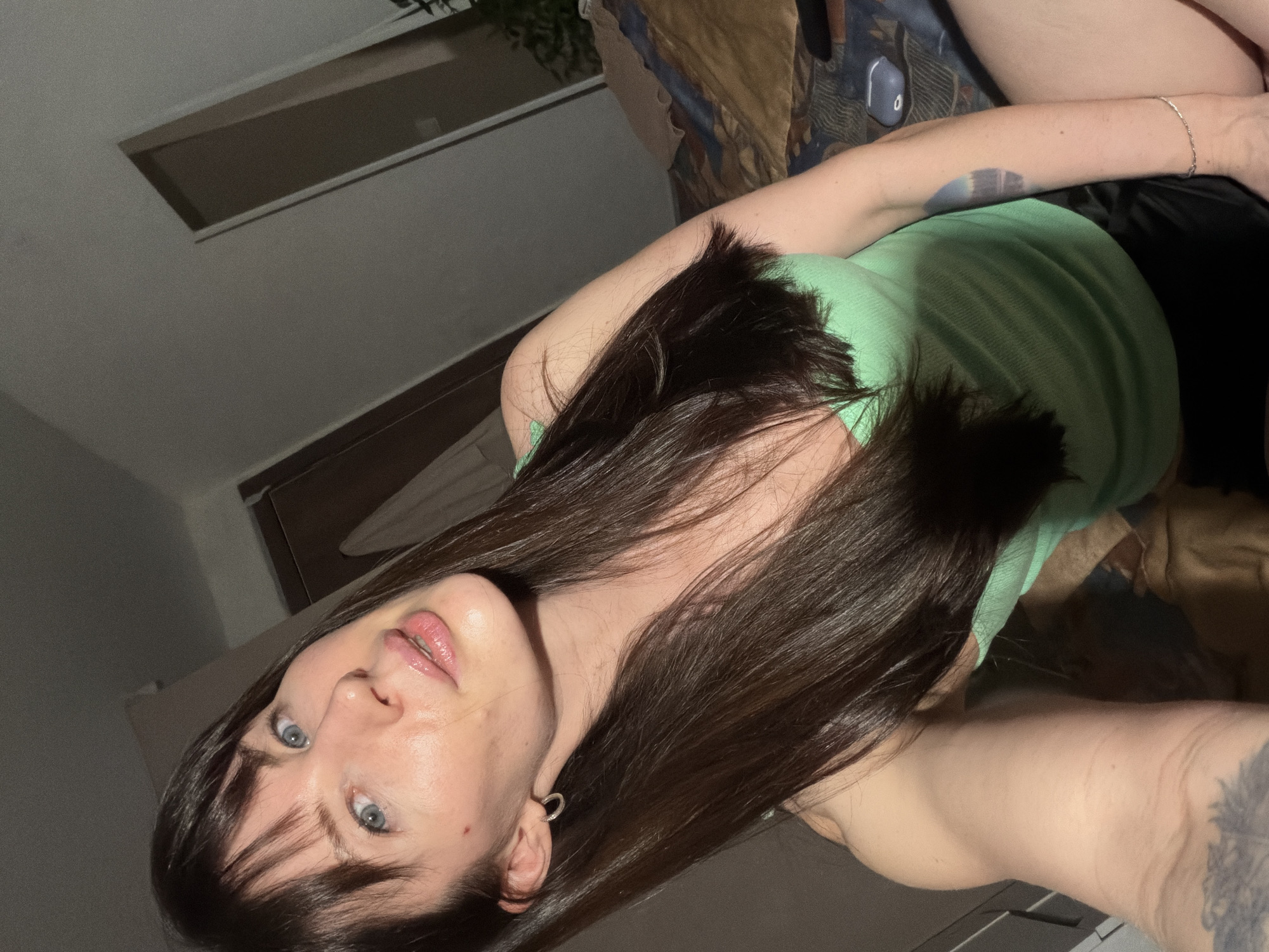 A person with long, dark hair, blue eyes, and fair skin is lying on a bed. They are wearing a light green tank top and dark shorts. The person has a tattoo on their left forearm and an earring in their left ear. The background includes a window, part of a bed frame, and a patterned blanket. The location is unknown and cannot be verified as Perm, Russia from this image.