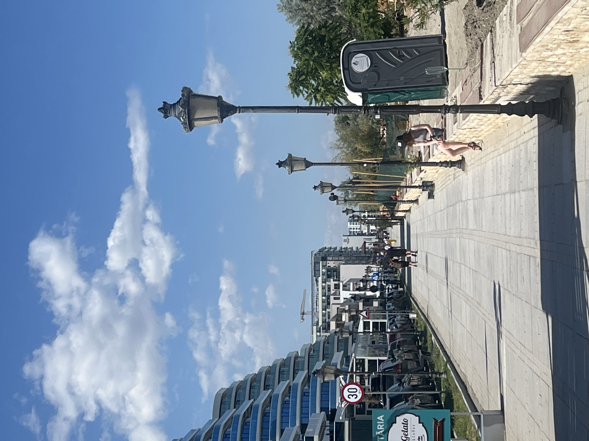This media file shows a street scene in Năvodari, Romania. The image features a paved walkway lined with lampposts, a portable toilet, and modern buildings with a curved architectural style. Several people are visible, some walking and one lounging on a bench. Cars are parked along the street.
The time of day appears to be daytime, with clear blue skies and scattered clouds indicating fair weather. The presence of a portable toilet and people enjoying the outdoors suggests a public area, possibly a boardwalk or promenade.
Notable details include the distinctive architecture of the buildings, the vintage style of the lampposts, and a "30" speed limit sign. There is also text visible on signs, including "TARIA" and "Gelato Coffee," suggesting nearby businesses. The overall atmosphere seems relaxed and casual, with people enjoying a sunny day.