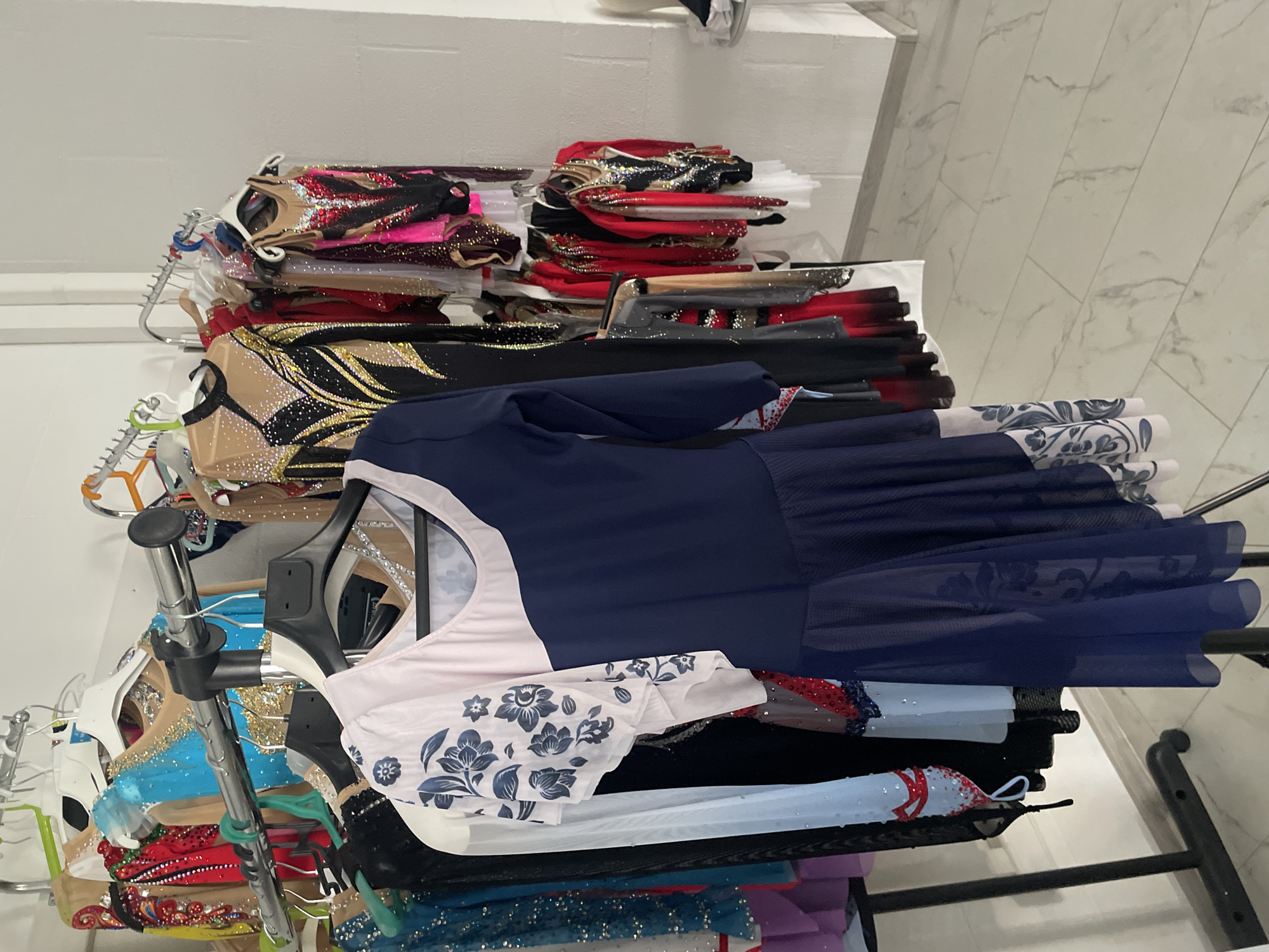 The image shows a rack of rhythmic gymnastics leotards hanging in a room with white walls and a light-colored tiled floor. The leotards are varied in color and design, many of them embellished with sequins and intricate patterns. Some are long-sleeved, while others are sleeveless. In the foreground, a dark blue leotard with a light pink floral-patterned sleeve is prominently displayed. The leotards are on hangers and appear to be organized on a metal garment rack. The overall scene suggests a storage or preparation area for a gymnastics team or individual athlete. There are no people visible in the image. The location is identified as Saint Petersburg, Russia.