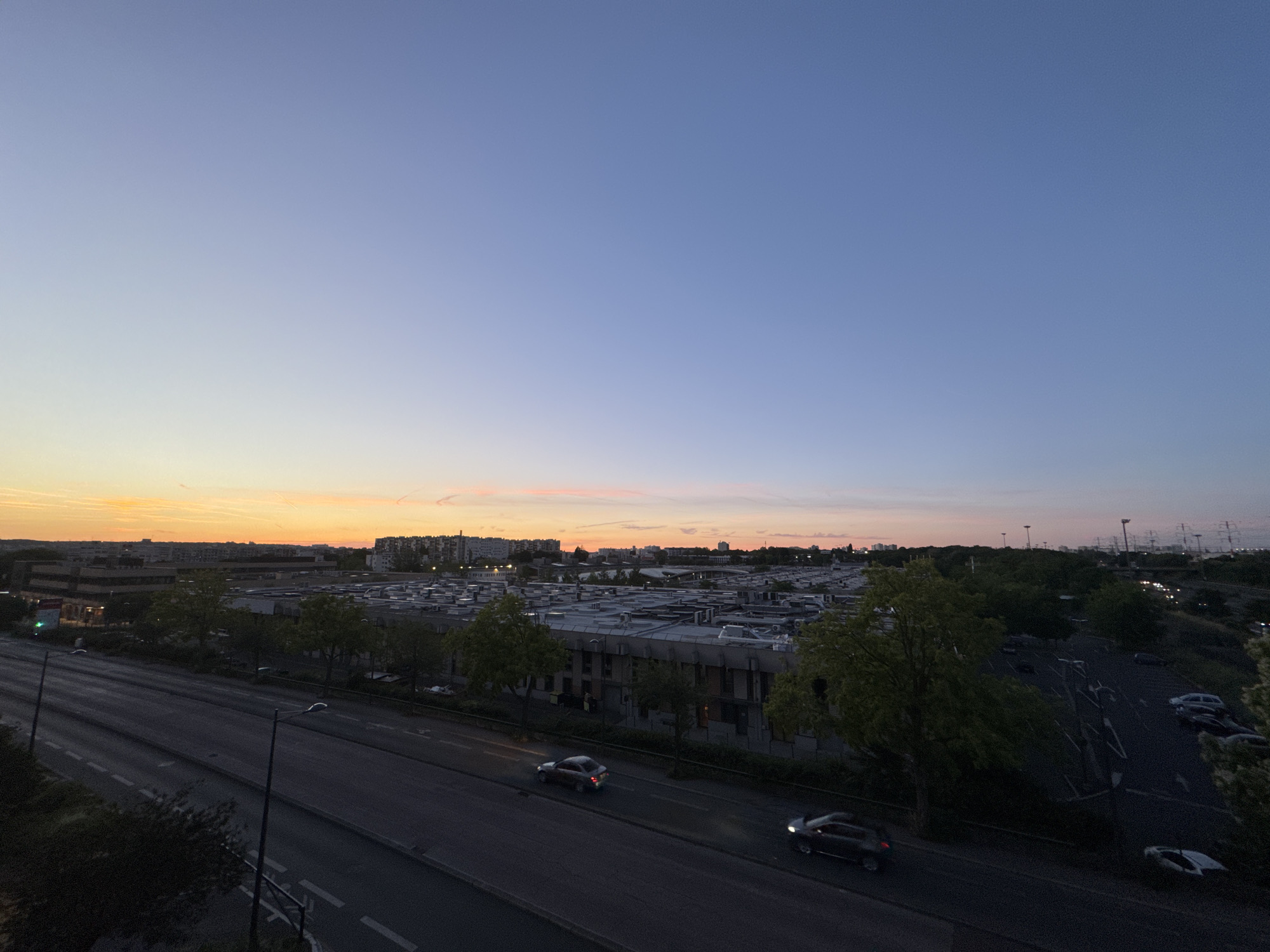 The image captures a wide-angle view of a city scene at dusk, with the sky transitioning from a pale orange at the horizon to a deep blue overhead. In the foreground, a multi-lane road runs diagonally from the bottom left towards the middle right of the frame, with several cars visible on it. One car, a silver sedan, is in motion in the left lane, while a dark sedan is also moving in the right lane further down the road. Streetlights are present along the road.
Behind the road, a low-rise industrial or commercial building complex stretches across the mid-ground, characterized by its flat roofs and numerous air conditioning units or vents. Trees with green foliage are interspersed between the road and the buildings, and also within the complex itself.
In the background, more urban structures and apartment buildings are visible, forming a dense skyline. The far background features a hazy horizon with faint lights, suggesting a sprawling metropolitan area. The overall impression is one of a typical urban periphery during the evening transition from day to night. The scene appears to be set in a location that is not immediately identifiable, but suggests a developed urban environment.