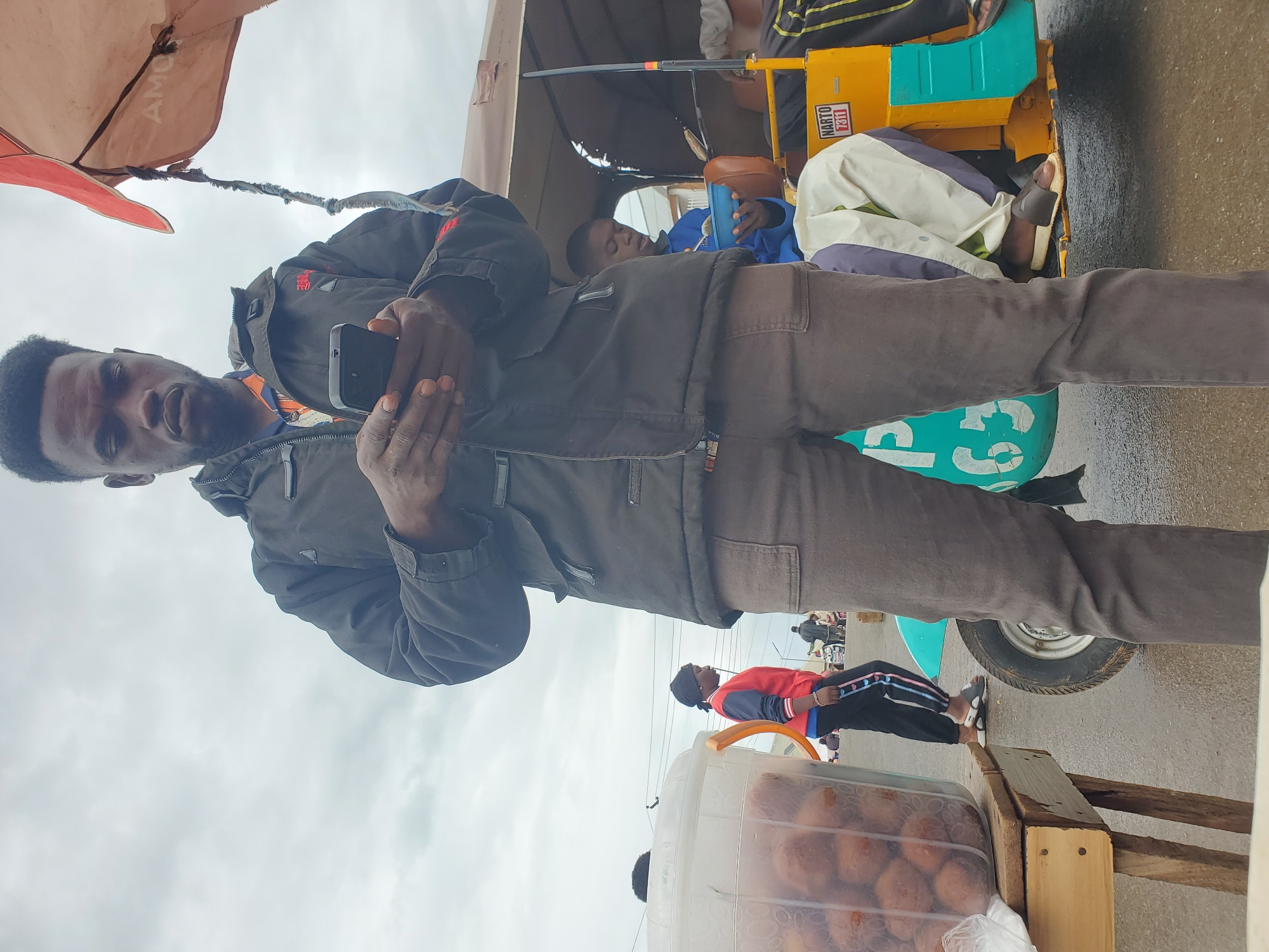 A medium shot captures a Black male, center frame, looking down at his black smartphone, held in both hands. He is wearing a dark jacket over an orange and black patterned shirt, and brown pants. To his left, a clear plastic container on a wooden stand is filled with brown spherical objects, possibly food. Behind him, a young Black female in a red and blue jacket and black pants walks away from the camera. In the background, a yellow and teal auto-rickshaw, identified with the text "NARTO 7311", is parked. A person in a blue shirt is seated inside the rickshaw, eating from a blue bowl. The scene appears to be an outdoor market or street in Gwong, Nigeria, under an overcast sky.