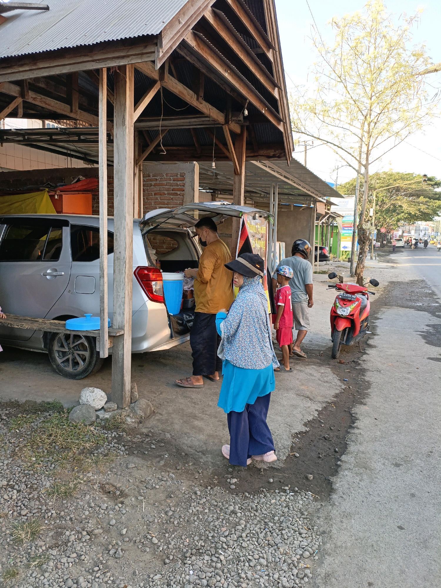 The image depicts a street scene in Bulukumba, Indonesia, likely near a roadside stall. In the foreground, a woman wearing a blue hijab and a patterned blue and white top stands on a gravel and asphalt surface. To her left, a silver minivan with its trunk open is parked. A wooden bench is placed in front of the minivan. Inside the trunk, there are blue containers and a person in an orange shirt wearing a black face mask is seen. A red scooter is parked on the asphalt to the right of the minivan. Further down the street, there are buildings, trees, and other vehicles, suggesting a commercial or residential area. The overall lighting suggests it is daytime.