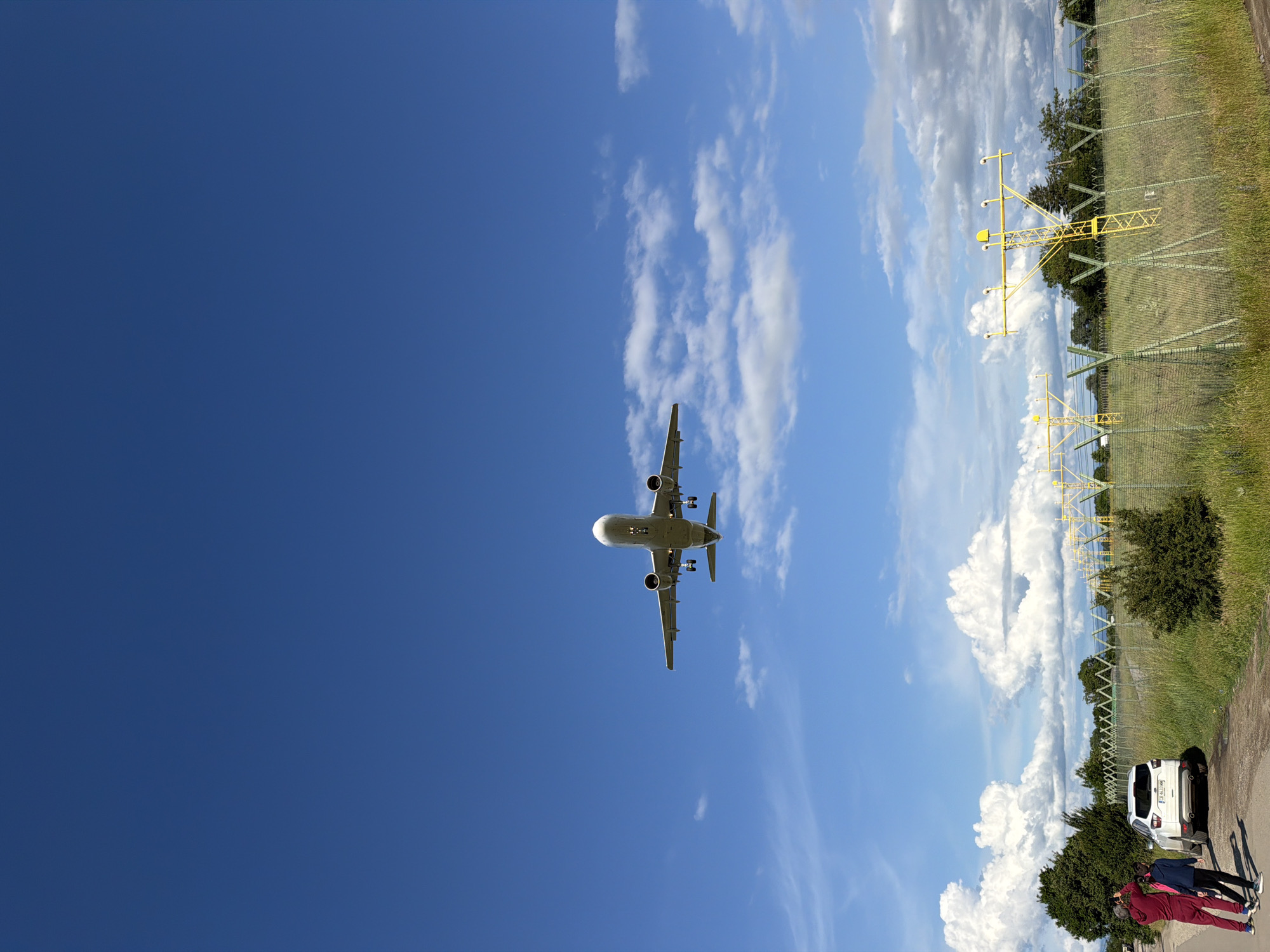 An airplane descends for landing against a predominantly deep blue sky, viewed from below. The aircraft, likely an Airbus A320 family type, is captured with its landing gear deployed, positioned towards the center-left of the frame. The sky transitions to a lighter blue with scattered white cumulus clouds on the right side of the image.
On the far right, at ground level, two individuals are visible, standing on a paved area. One person wears red pants and a dark top, while the other is dressed in a dark jacket. They appear to be looking upwards, observing the airplane. A white SUV or hatchback is parked near them, with a partially visible Bulgarian license plate reading "CB 640_ AM".
Beyond the paved area, a green chain-link fence separates the foreground from a grassy field and trees. Several yellow metal structures, which are identifiable as airport approach or landing lights, stand tall behind the fence and foliage, indicating proximity to a runway.
The scene depicts a clear, sunny daytime environment, likely late morning or early afternoon, at an airport perimeter. The presence of the landing aircraft, airport lights, and the license plate suggests this is near Sofia Airport in Sofia, Bulgaria, with people engaging in plane-spotting.