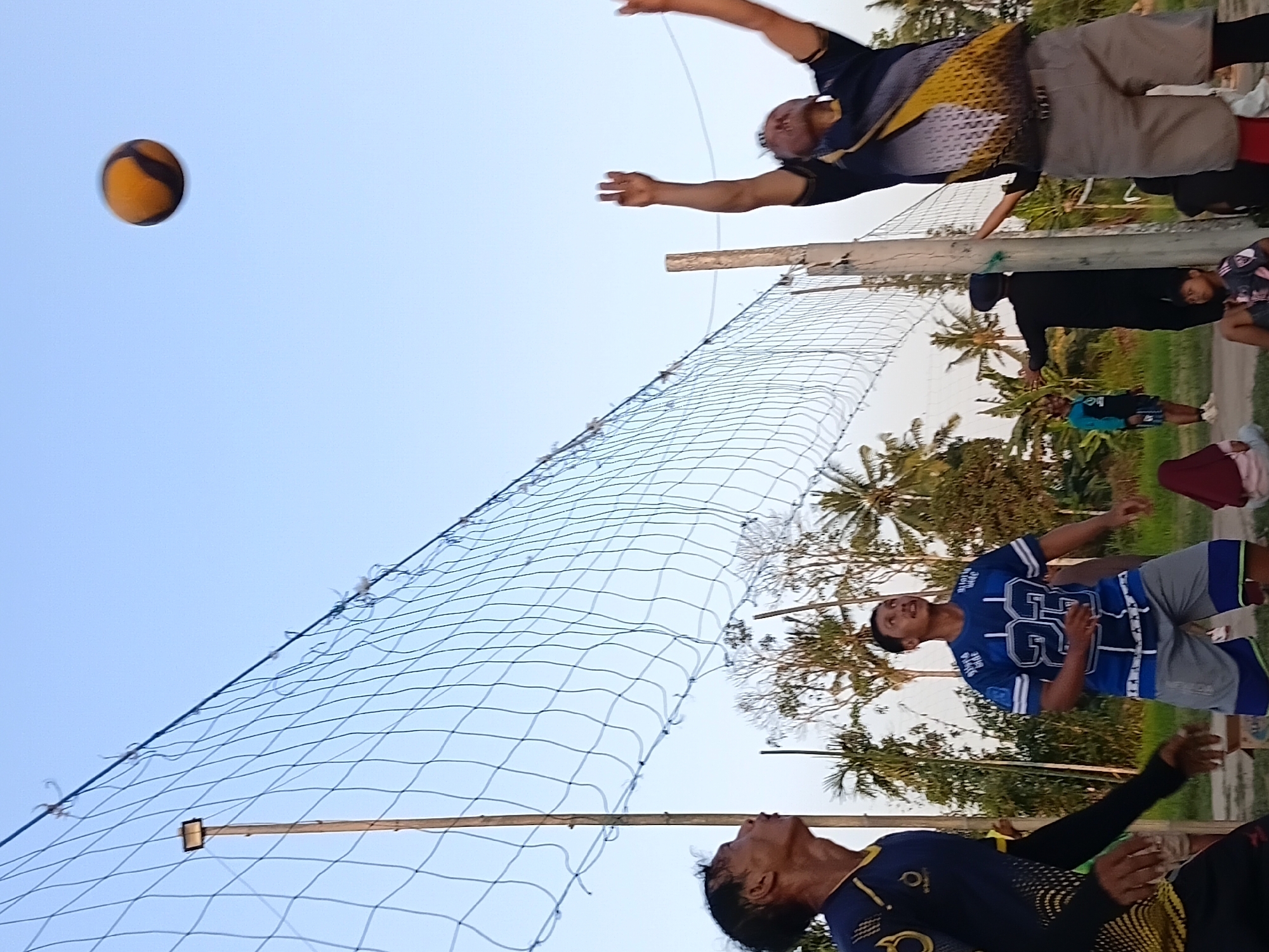 In Margo Rukun, Indonesia, a group of people are playing volleyball on a dirt court with a net stretched across it. The sky is clear and a light orange color, suggesting it is either sunrise or sunset. Two men are actively engaged in the game, one jumping to spike the ball, which is visible in the air, and another looking up at it. Another man in a blue jersey stands ready on the other side of the net. Several other people, including children and women, are present, some watching the game and others sitting. Palm trees and other vegetation are visible in the background, indicating a rural or suburban setting.