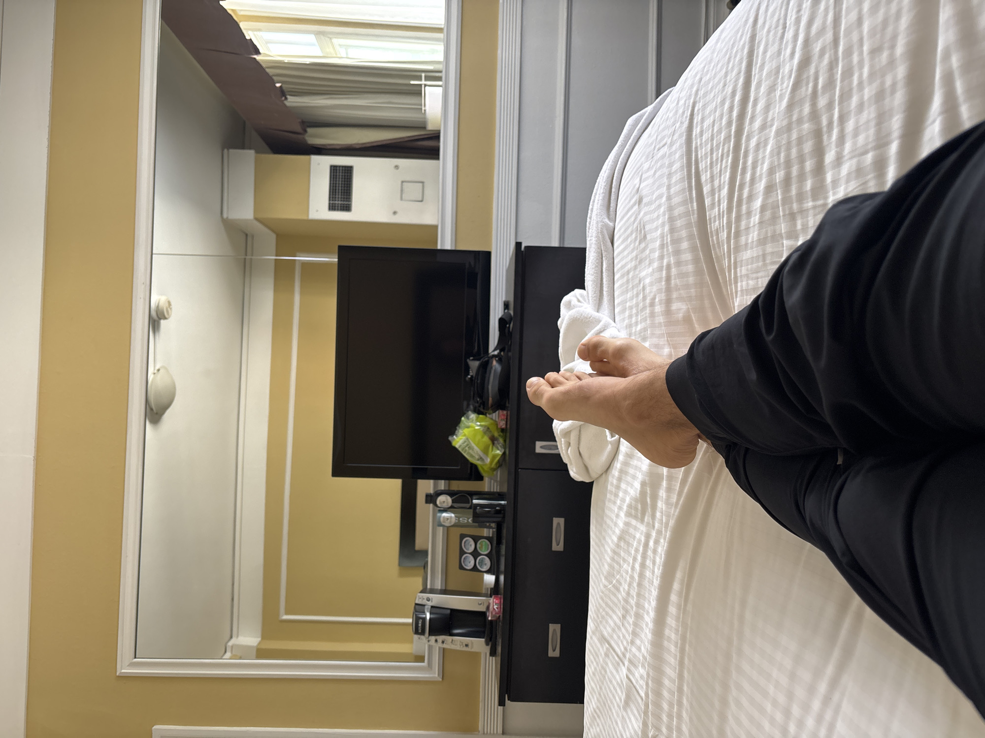 This is a first-person perspective shot of a person's feet and legs resting on a white, striped bed in a hotel room. The person is wearing black pants. In the background, there is a dresser with a television and a Keurig coffee maker. A large mirror hangs above the dresser, reflecting the room, including a window with curtains and an air conditioning unit. The walls are painted a pale yellow. The location is not specified.
