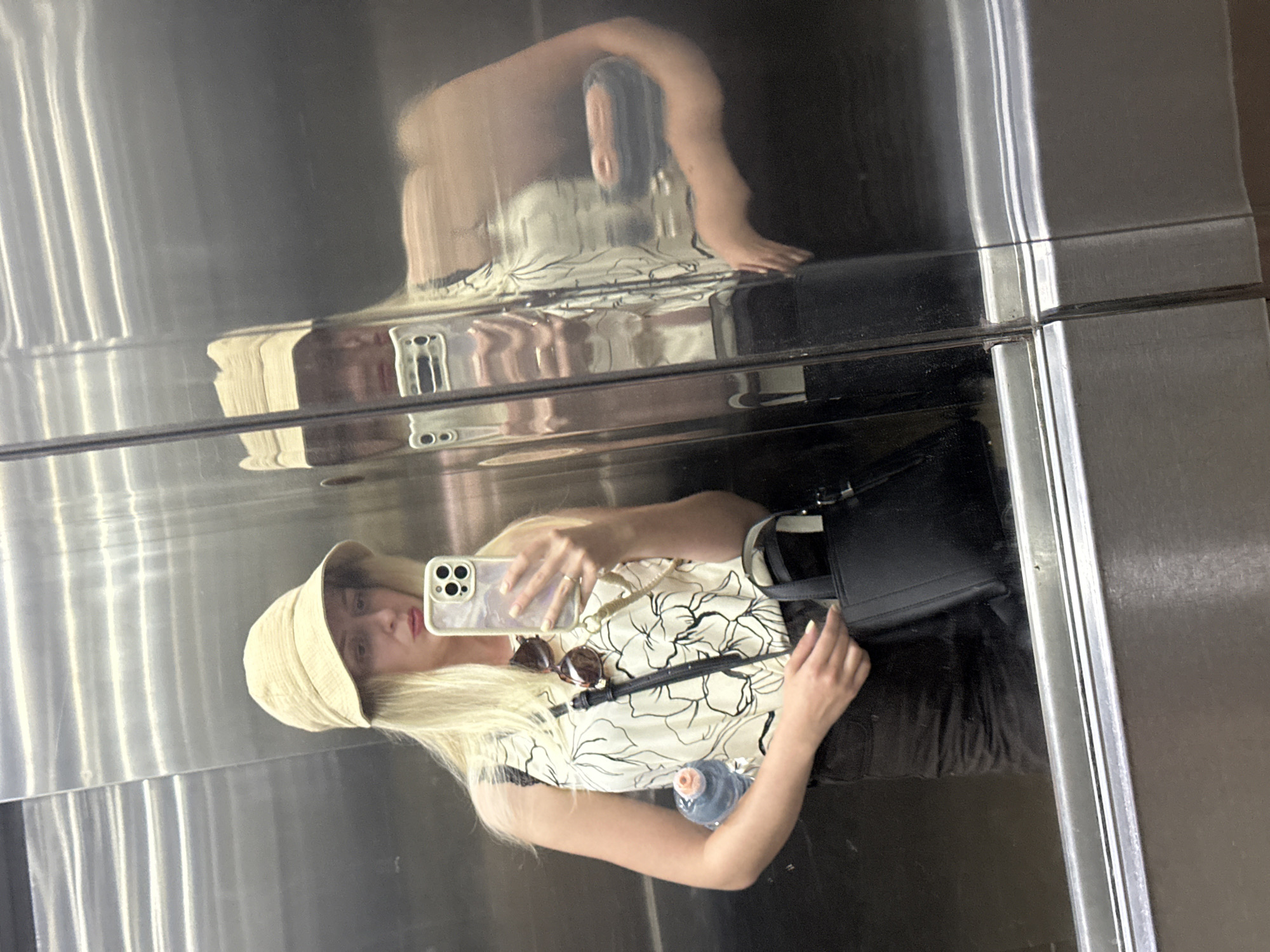 The image depicts a woman taking a mirror selfie inside an elevator, located in Petah Tikva, Israel. The woman, with long blond hair, is wearing a light beige bucket hat and a cream-colored sleeveless top featuring a black floral pattern. She is also wearing dark-colored bottoms, possibly shorts or a skirt, with a slight sheen. Dark-lensed sunglasses are hooked onto the neckline of her top.
In her right hand, she holds a light-colored smartphone with a distinctive multi-lens camera array, housed in a light, possibly iridescent, case. Her left hand holds a small blue water bottle with a pink cap and also grips the strap of a small, structured black handbag. A gold-toned ring is visible on the ring finger of her left hand.
The elevator interior features highly polished, silver-toned metallic walls with visible horizontal panel lines. A distinct vertical seam divides the reflective surface near the center, creating a slightly distorted reflection of the woman on the right side of the frame. A horizontal metallic railing or trim is visible towards the bottom of the elevator wall.