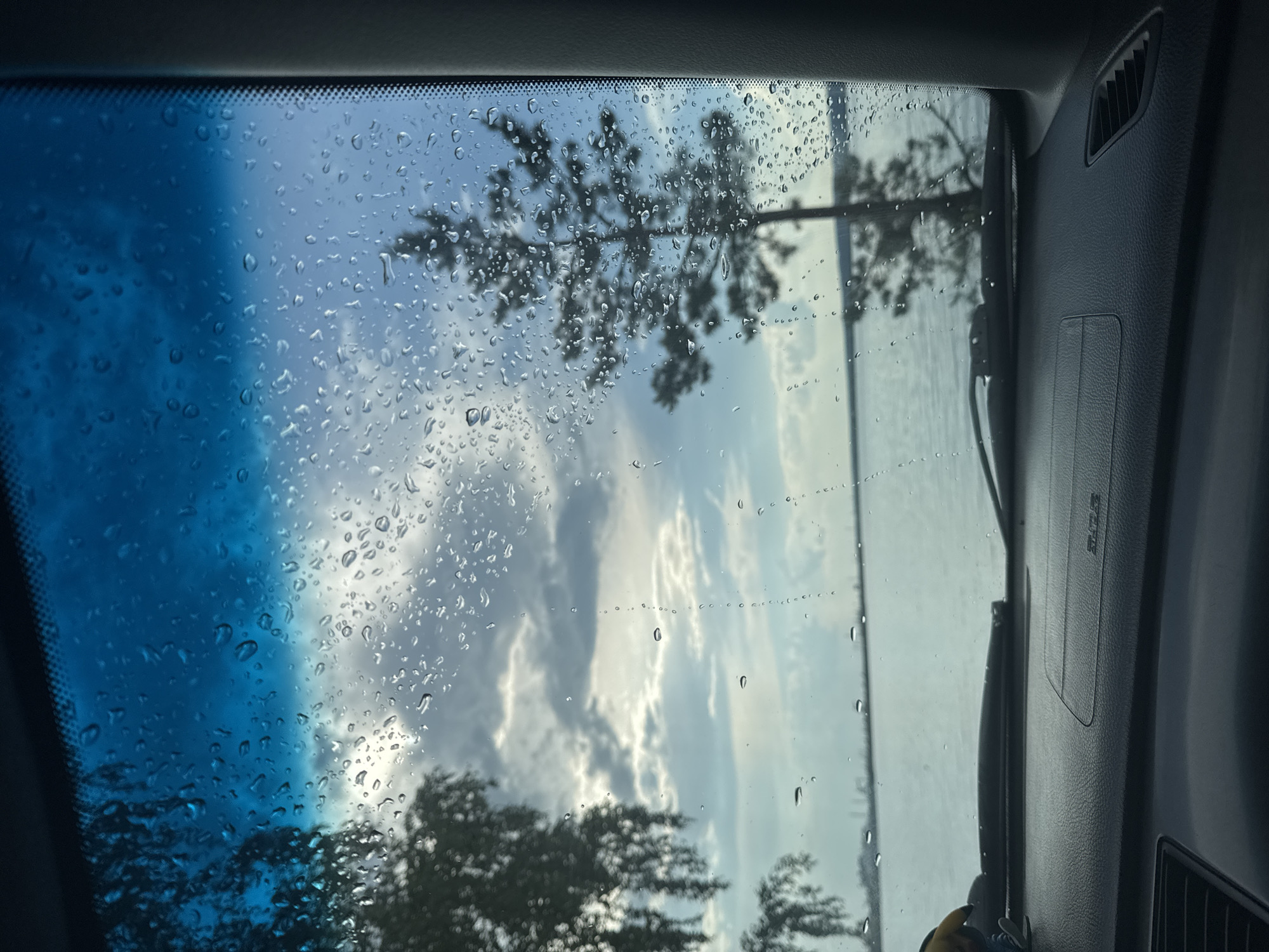 The image is taken from inside a vehicle, looking out through a rain-streaked windshield at a body of water, likely a river or reservoir, on a cloudy day. The foreground shows the dashboard of the car, with a small yellow toy figure on the left. In the middle ground, a calm body of water stretches to the horizon, with distant structures visible. On the left side of the frame, a silhouette of a leafy tree is visible against the sky. On the right side, a tall, slender pine tree is silhouetted against the sky. The sky is a mix of blue and gray clouds, with sunlight filtering through in places, creating bright patches. The overall mood is serene and contemplative. The location is identified as Chaikovsky, Russia, likely a view of the Kama River reservoir, which is a prominent feature of the city.