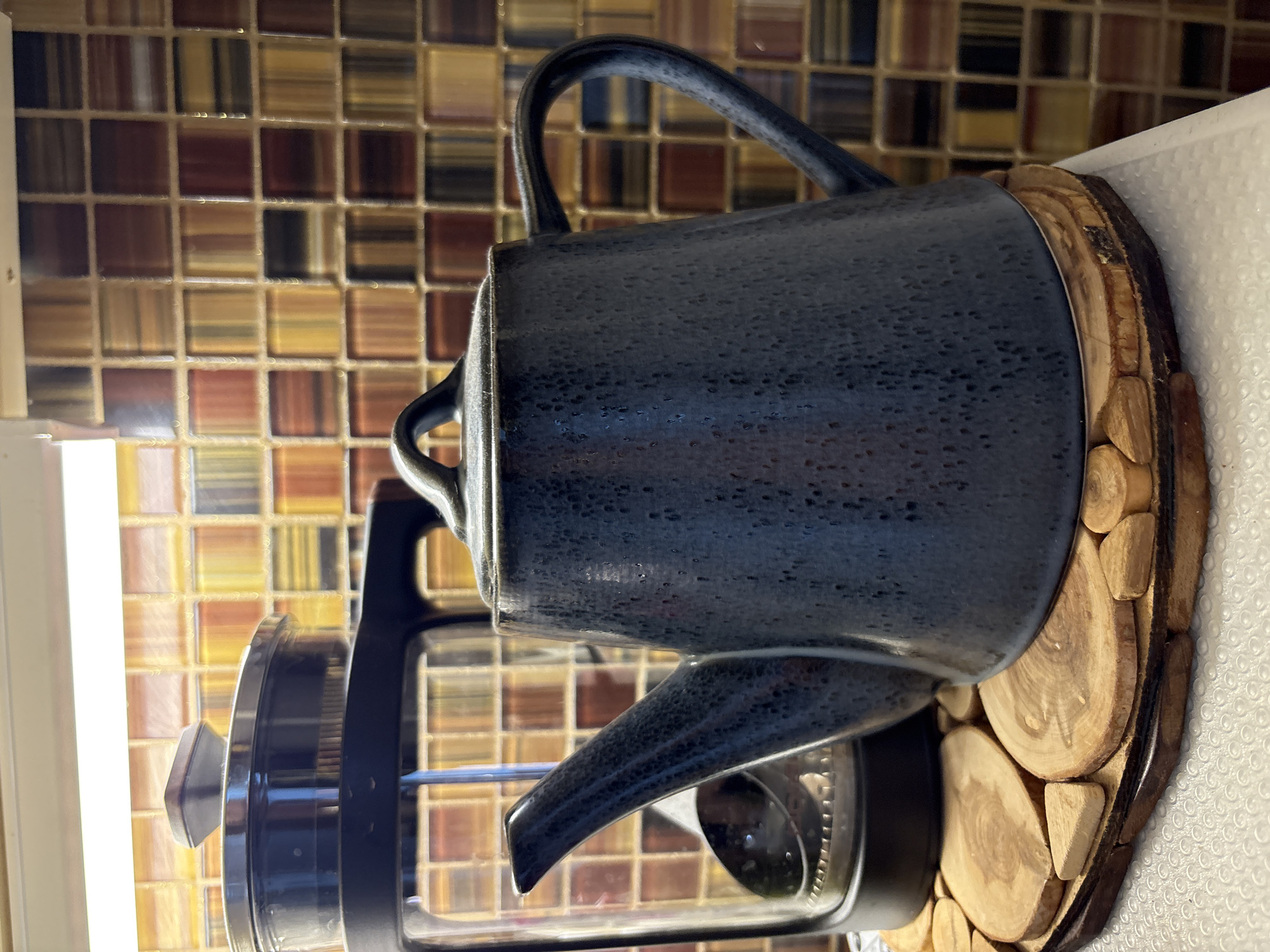 A close-up shot of a dark blue ceramic kettle placed on a wooden trivet. The kettle is situated next to a French press coffee maker. The backdrop consists of a multicolored tiled wall. The location is not discernible from the provided image, and therefore cannot be pinpointed to Chaikovsky, Russia.