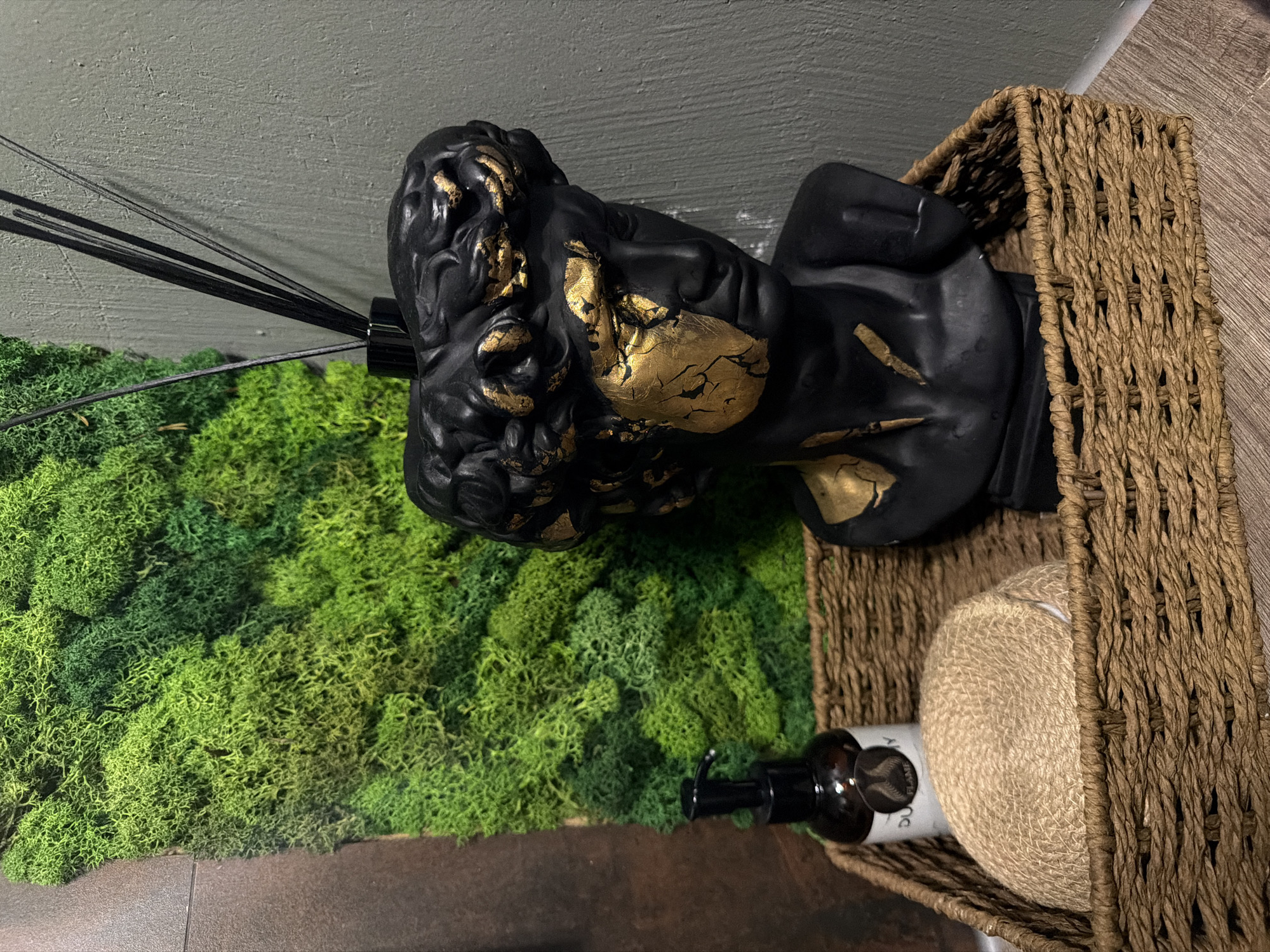 This is a close-up, eye-level, indoor shot of a decorative arrangement on a shelf or counter. The background features a textured, grey wall and a vertical panel of bright green preserved moss. In the foreground, a woven brown basket holds several items. Prominently displayed is a black bust of a head with gold accents, resembling classical sculptures. To the left of the bust, inside the basket, is a small amber-colored bottle with a black pump dispenser, and a round, tightly wound coil of natural fiber, possibly for bathing or cleaning. Behind the bust, several thin black diffuser reeds extend upwards from a black base. The scene is well-lit, highlighting the textures and colors of the objects.