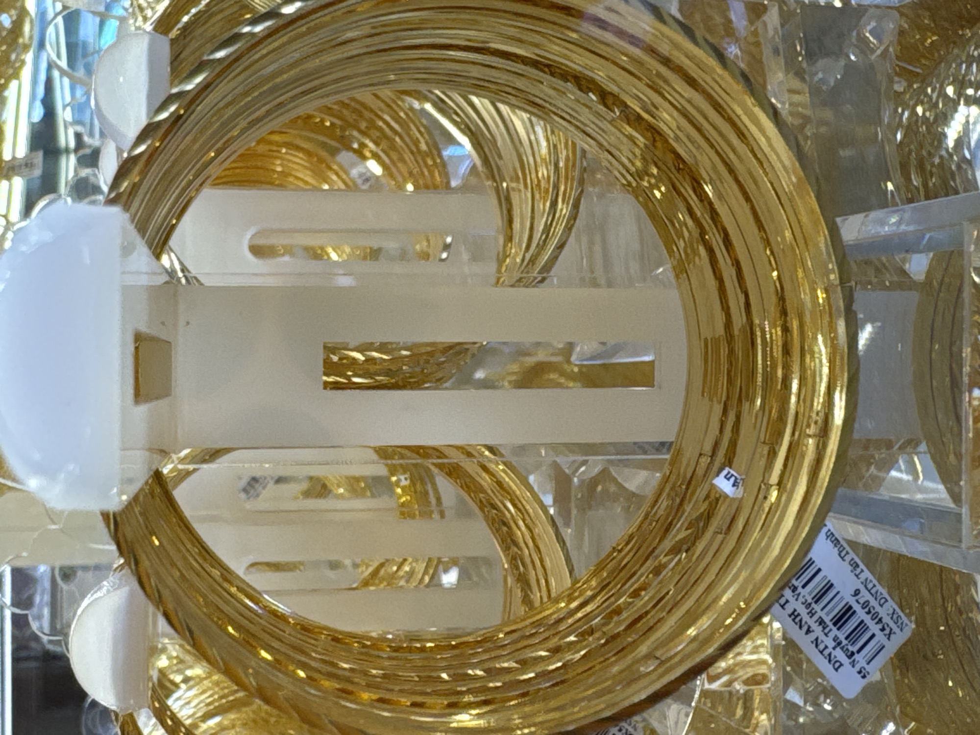 The image shows a close-up view of numerous gold-colored bangles displayed in a store. The bangles are stacked and arranged on white plastic stands. Some of the bangles appear to be individually packaged in clear plastic. A label with a barcode and text in Vietnamese is visible on one of the bangles. The lighting suggests an indoor retail environment, likely a jewelry shop. The text on the label indicates "DNTN ANH TI", "55 Nguyễn Thái Học, Var", "X5405076", and "NSX: DNTN Tân Thành". The location in Nha Trang Ward, Vietnam, is inferred from the provided context, suggesting this is a retail establishment in that area. No people are visible in the image.