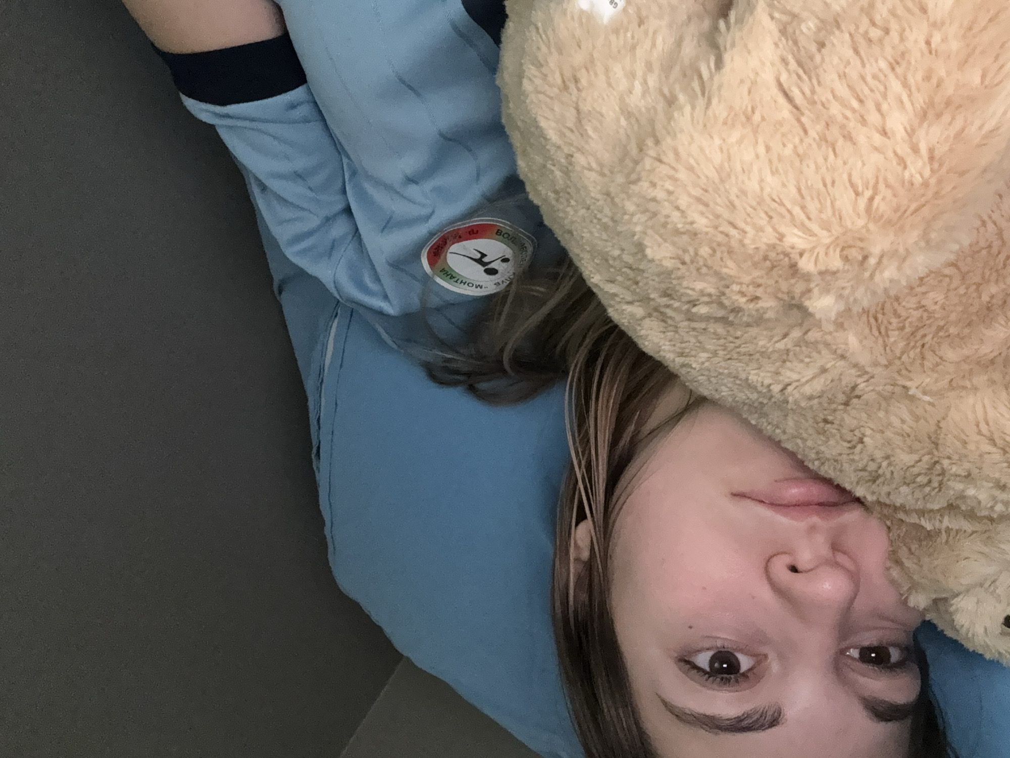 A person is lying down, face visible, with a beige plush object partially covering the face. The person has light brown hair and is wearing a light blue garment with a patch that includes the words "МОНТАНА" and "ВОЛЖ" with an image of a diver. The background includes a blue pillow, a gray surface, and a dark blue sleeve. The location in the city of Sofia, Bulgaria cannot be determined from this image.