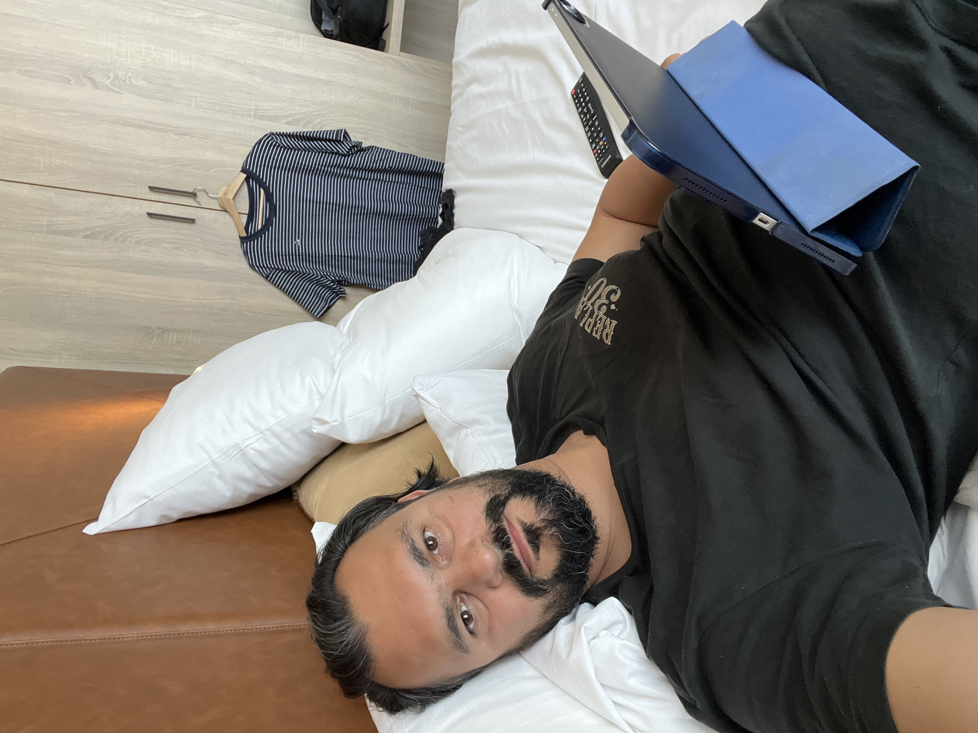 A person is lying on a bed in what appears to be a hotel room in Delft, Netherlands. The room has light-colored wooden paneling on the wall and a brown leather headboard. The person is wearing a black t-shirt with text on it that reads "REPLICA 20". They are holding a blue tablet or laptop. Next to the person, on the bed, are several white pillows and a remote control. On the floor, a striped t-shirt is hanging on a clothes hanger. The lighting suggests it is daytime, possibly late afternoon or evening, as there is no direct sunlight. The overall mood is relaxed and casual, as the person is comfortably lying on the bed.