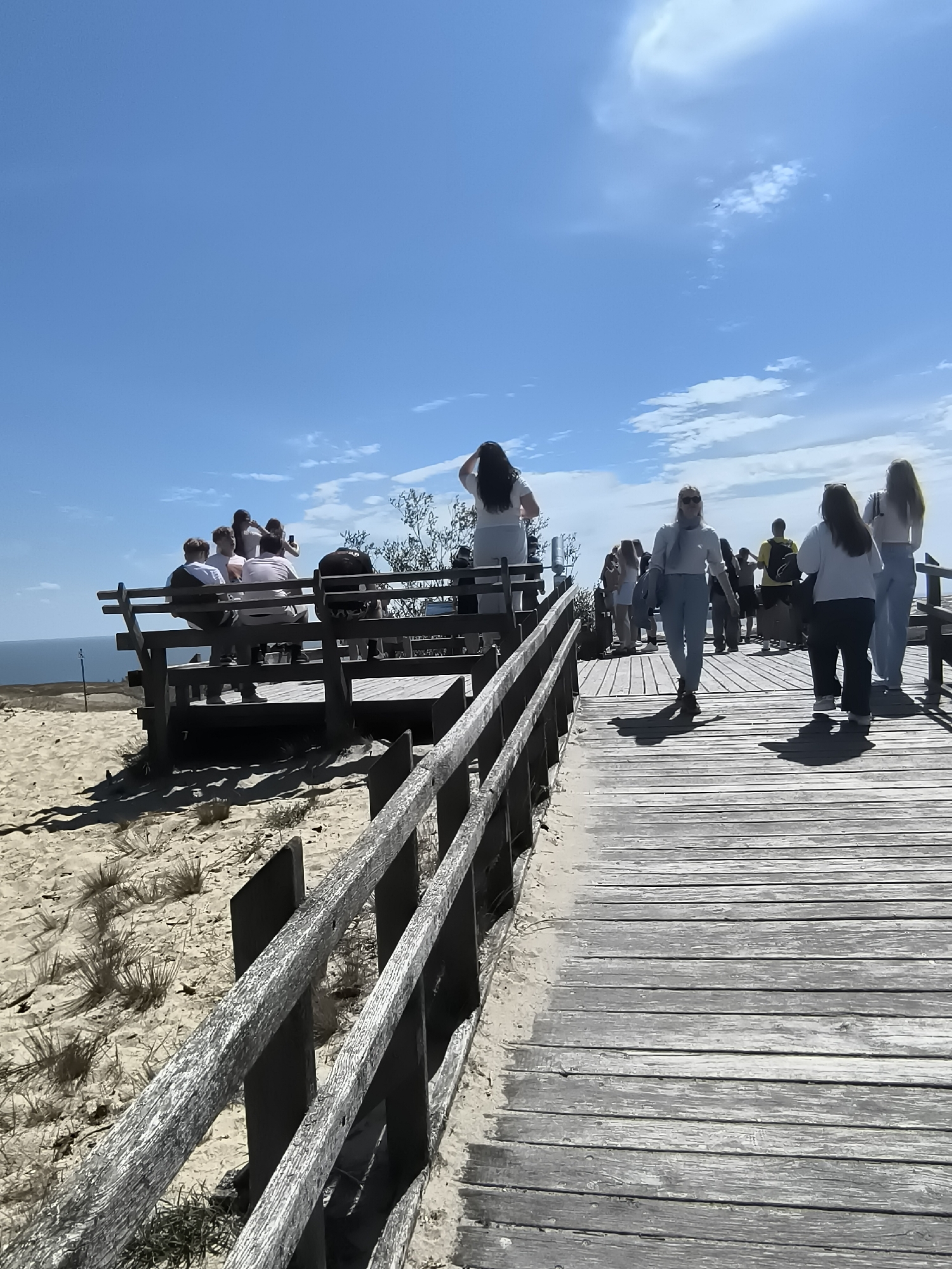 The image depicts a sunny day with a clear blue sky and scattered clouds, possibly at the Curonian Spit in Neringa, Lithuania, given the sandy terrain and boardwalk. A wooden boardwalk stretches diagonally across the frame, leading towards a raised wooden viewing platform. Several people are present on the boardwalk and the platform. On the platform, a group of individuals is gathered, some seated and others standing, appearing to be enjoying the view or taking pictures. On the boardwalk, individuals are walking in various directions, suggesting a recreational area. The environment is characterized by sand dunes, sparse vegetation, and a glimpse of the sea or a large body of water in the distance. The wooden structures, both the boardwalk and the platform, appear weathered, indicating exposure to the elements. The overall scene conveys a sense of a popular tourist or recreational spot.