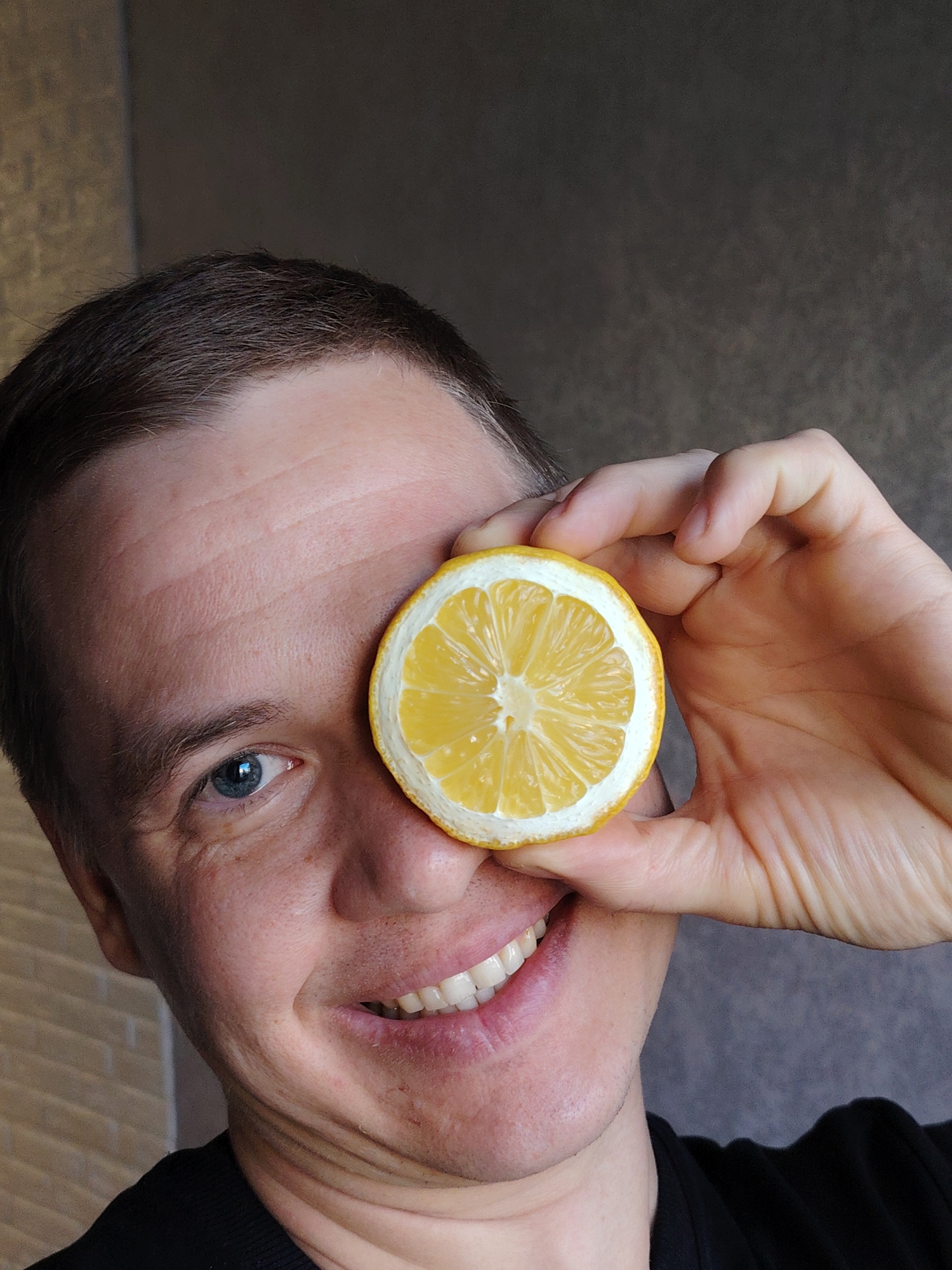 A smiling man with short brown hair and blue eyes is holding a slice of lemon over his right eye. The man is wearing a black shirt. The background appears to be a wall with a subtle texture. There is no information to determine the location in the city of Chaikovsky, Russia.