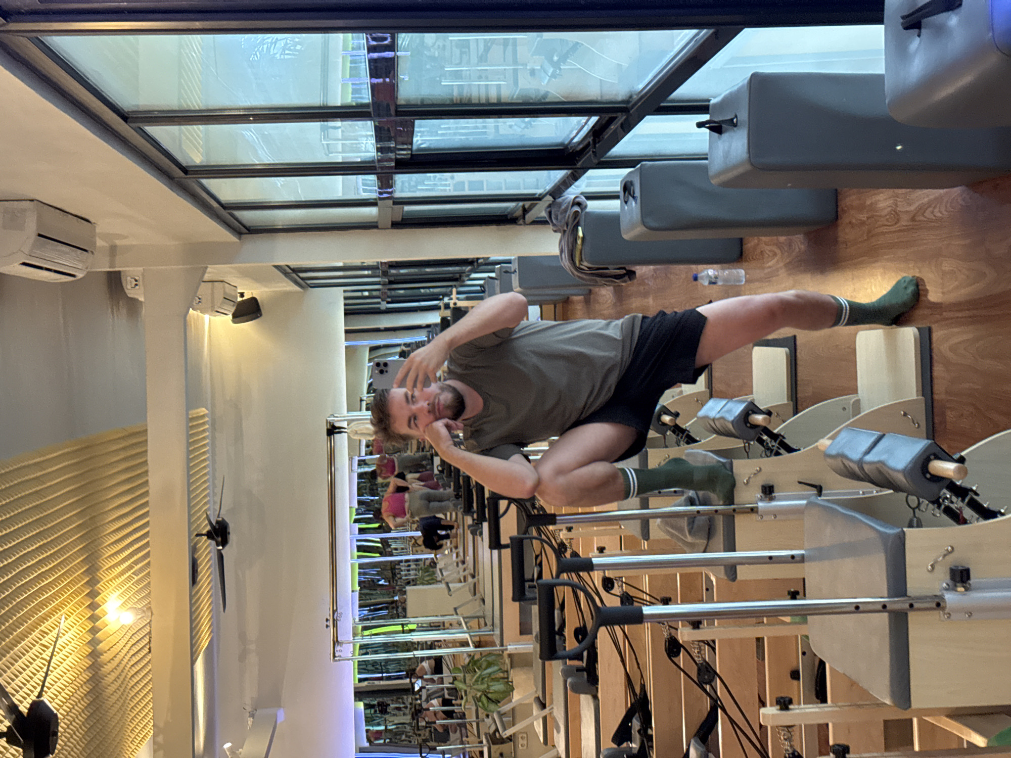 The image captures an indoor Pilates studio in North Kuta, Indonesia. A light-skinned man with a beard, wearing a green t-shirt, black shorts, and green socks with white stripes, is the central subject. He is lying on his back on a light wooden Pilates reformer, propped up on his left elbow with his hand resting on his chin, holding a smartphone in his right hand, and looking at the camera with a slight smile. His left leg is extended, and his right leg is bent.
Multiple other Pilates reformers, also made of light wood with grey padded sections, are visible throughout the studio, some extending into the foreground and background. A clear plastic water bottle is on the wooden floor near the man's extended leg. High on the wall, an air conditioning unit is mounted, and above it, a large glass skylight forms part of the ceiling, contributing to the bright illumination. Two dark-bladed ceiling fans are positioned on the left wall, alongside a large mirror that reflects other individuals, mostly women, and more of the studio, suggesting a group class is in session. A ribbed, light-colored wall panel is visible on the far left.
The man appears relaxed and is playfully posing for a photo or selfie amidst his workout. The studio features light wooden flooring and light-colored walls, creating an open and clean aesthetic. No discernible text is visible in the image.
