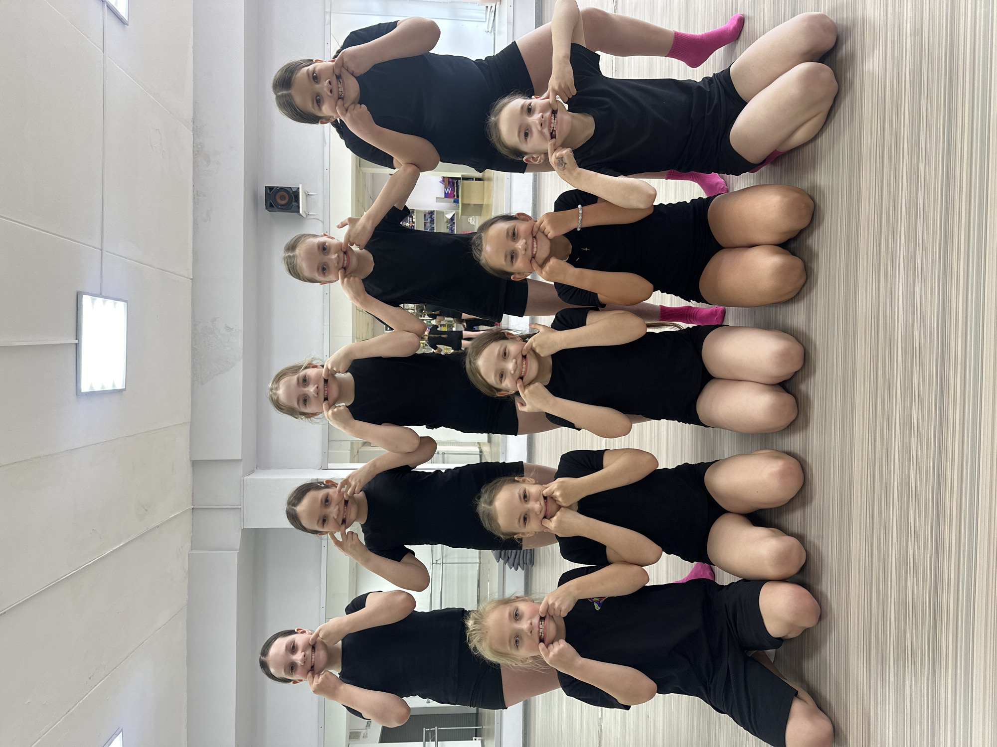 Eight young girls are captured in a playful indoor group photo, likely taken in a dance or gymnastics studio in Votkinsk, Russia. The girls are arranged in a horizontal pyramid-like formation, lying on their stomachs, one on top of the other, with their heads facing the camera. Each girl is making a humorous "silly face" by pulling at her cheeks with her fingers, revealing her teeth and gums in a wide, exaggerated smile.
They are uniformly dressed in black athletic short-sleeved tops and matching black shorts. Several of the girls add a pop of color with bright pink socks. The setting features light-colored, vertically striped wooden floorboards and light-toned walls with rectangular ceiling lights. A black speaker is mounted on the far wall. The overall mood of the image is joyful and lighthearted, capturing a moment of camaraderie and fun among the young participants. No discernible text is visible in the frame.