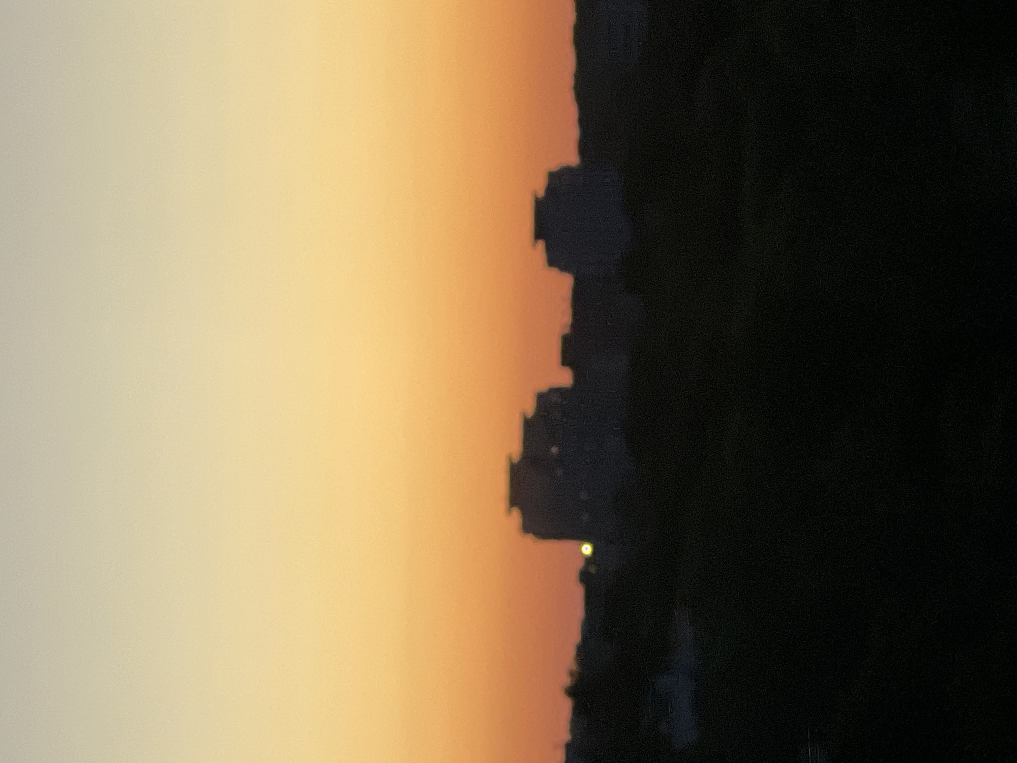 The image depicts a silhouette of a city skyline at dusk, with buildings and trees visible against a gradient of orange and pale yellow sky. The scene is dominated by dark, indistinct shapes of architectural structures and foliage. The sky transitions from a brighter yellow near the horizon to a lighter shade of yellow or cream at the top of the frame. The overall impression is one of a quiet evening in an urban environment.
Based on visual cues in the original image, the location is likely the city of Minsk, Belarus, specifically the area near the "Minsk World" residential complex or other similar high-rise developments. The architectural style of the silhouetted buildings, characterized by their stepped roofs and blocky forms, is consistent with modern construction in Minsk. While no specific landmarks are clearly identifiable, the general urban density and architectural typology point towards this region.