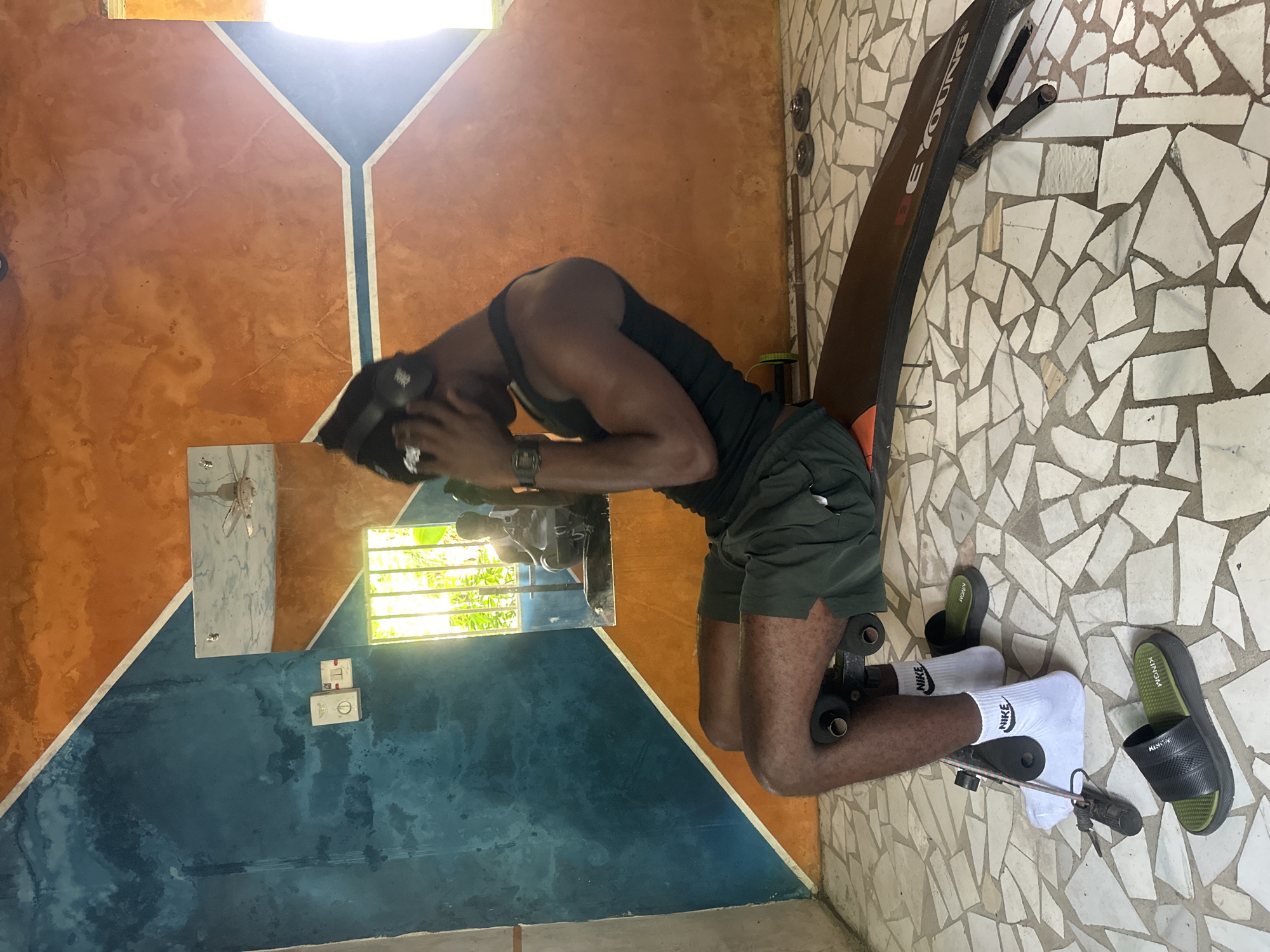 In Badagry, Nigeria, a person is performing sit-ups on a black weight bench in a room with a distinctive mosaic tiled floor. The walls are painted in bold geometric patterns of orange and teal. Natural light streams in from a window, illuminating the scene and casting shadows. The individual is wearing a black tank top, green shorts, white Nike socks, and black and green slides. They have headphones on and are holding their hands near their head. A mirror on the wall reflects another person sitting in the room. In the background, gym equipment, including weights and a resistance band, is visible on the floor.