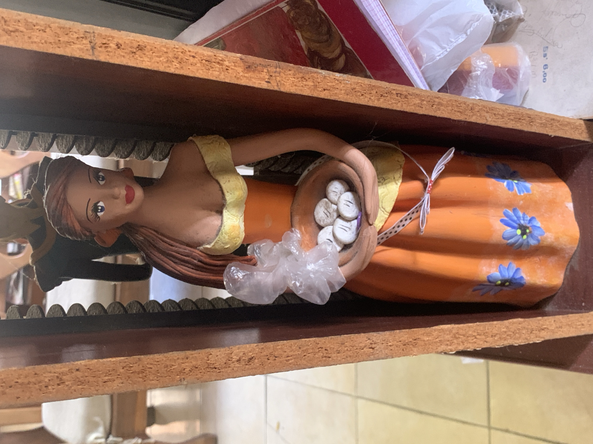 The image shows a ceramic figure of a woman with brown hair, an orange dress decorated with blue flowers, and a yellow sash. She is holding a basket containing round, white objects marked with lines. The figure is situated within a wooden shelf or display case with decorative trim. The background contains other items, including a wrapped object and paperwork. There is no confirmed geographic location within Santa Ana, Venezuela in this image.